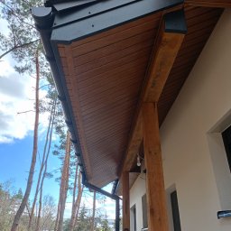 BudZan Usługi budowlane wykończeniowe domów, mieszkań oraz elewacje. - Drewniana podbitka dachowa z czarnymi rynnami, widok z dołu na tle błękitnego nieba i drzew. Elementy wykończeniowe elewacji domu jednorodzinnego.