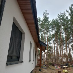 BudZan Usługi budowlane wykończeniowe domów, mieszkań oraz elewacje. - Biała elewacja domu z szarymi roletami i drewnianą podbitką, widok z boku na tle lasu. Świeżo otynkowana ściana z widocznymi detalami wykończenia.