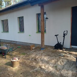 BudZan Usługi budowlane wykończeniowe domów, mieszkań oraz elewacje. - Biała elewacja domu po tynkowaniu, okna zabezpieczone folią, widoczny rower stacjonarny i taczka z narzędziami. Ujęcie z boku budynku, w tle las.