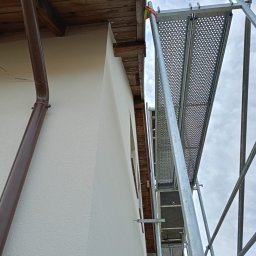 BudZan Usługi budowlane wykończeniowe domów, mieszkań oraz elewacje. - Świeżo otynkowana elewacja domu z brązową rynną, widoczna z dołu, z metalowym rusztowaniem na tle pochmurnego nieba. Detal wykończenia.
