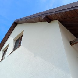 BudZan Usługi budowlane wykończeniowe domów, mieszkań oraz elewacje. - Biała elewacja domu jednorodzinnego z brązowym dachem i oknami, widok z dołu na tle błękitnego nieba. Tekstura tynku barankowego.