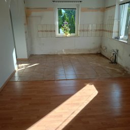 BudZan Usługi budowlane wykończeniowe domów, mieszkań oraz elewacje. - Pomieszczenie w trakcie remontu z podłogą łączącą jasne płytki i drewniane panele. Ściany z częściowo usuniętymi płytkami i fragmentami tynku, przygotowane do dalszych prac. Słońce tworzy ukośny cień.
