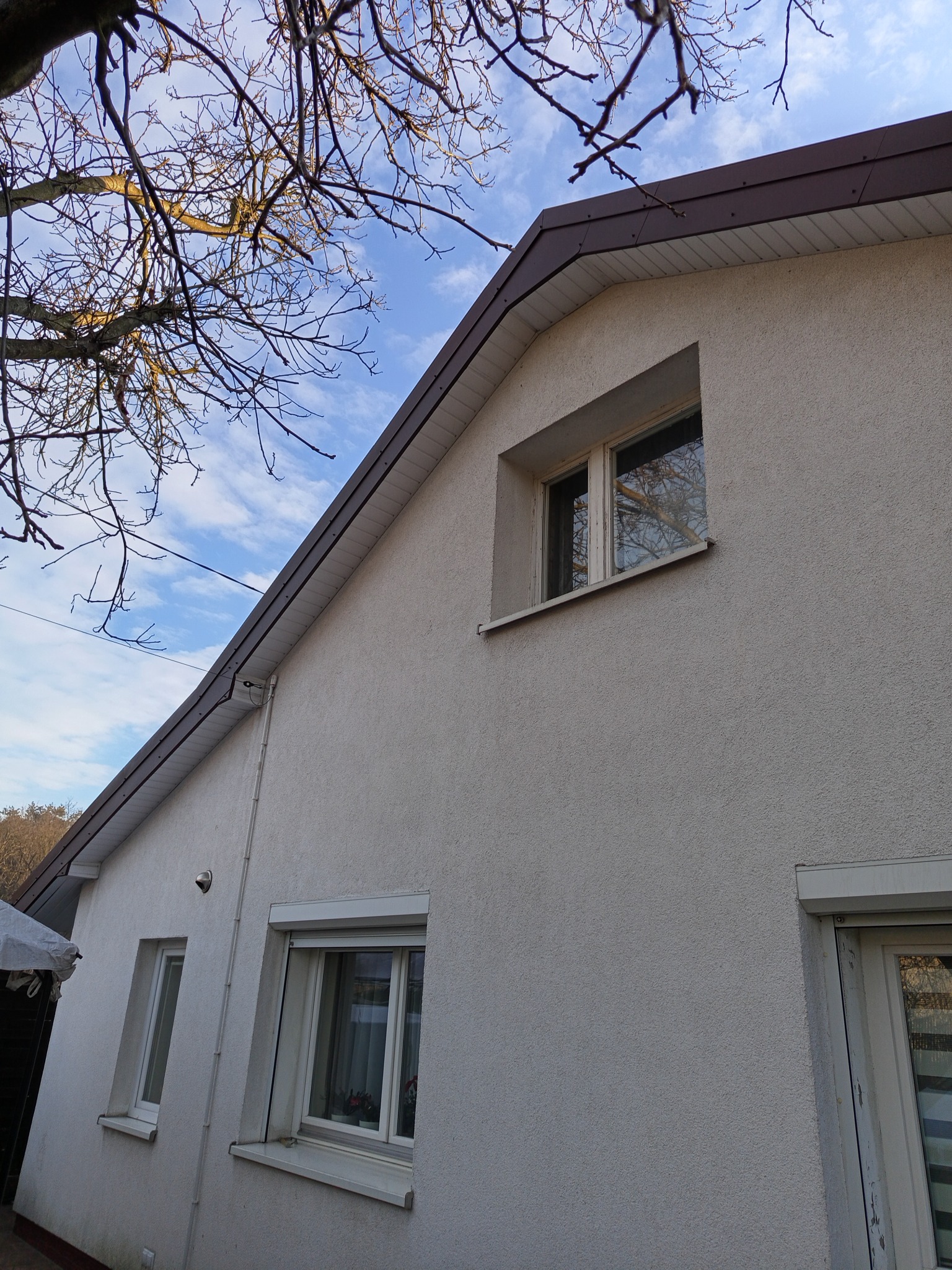 Elewacja domu jednorodzinnego z oknami i brązowym wykończeniem dachu, widziana pod kątem. Na pierwszym planie gałęzie drzewa na tle błękitnego nieba.