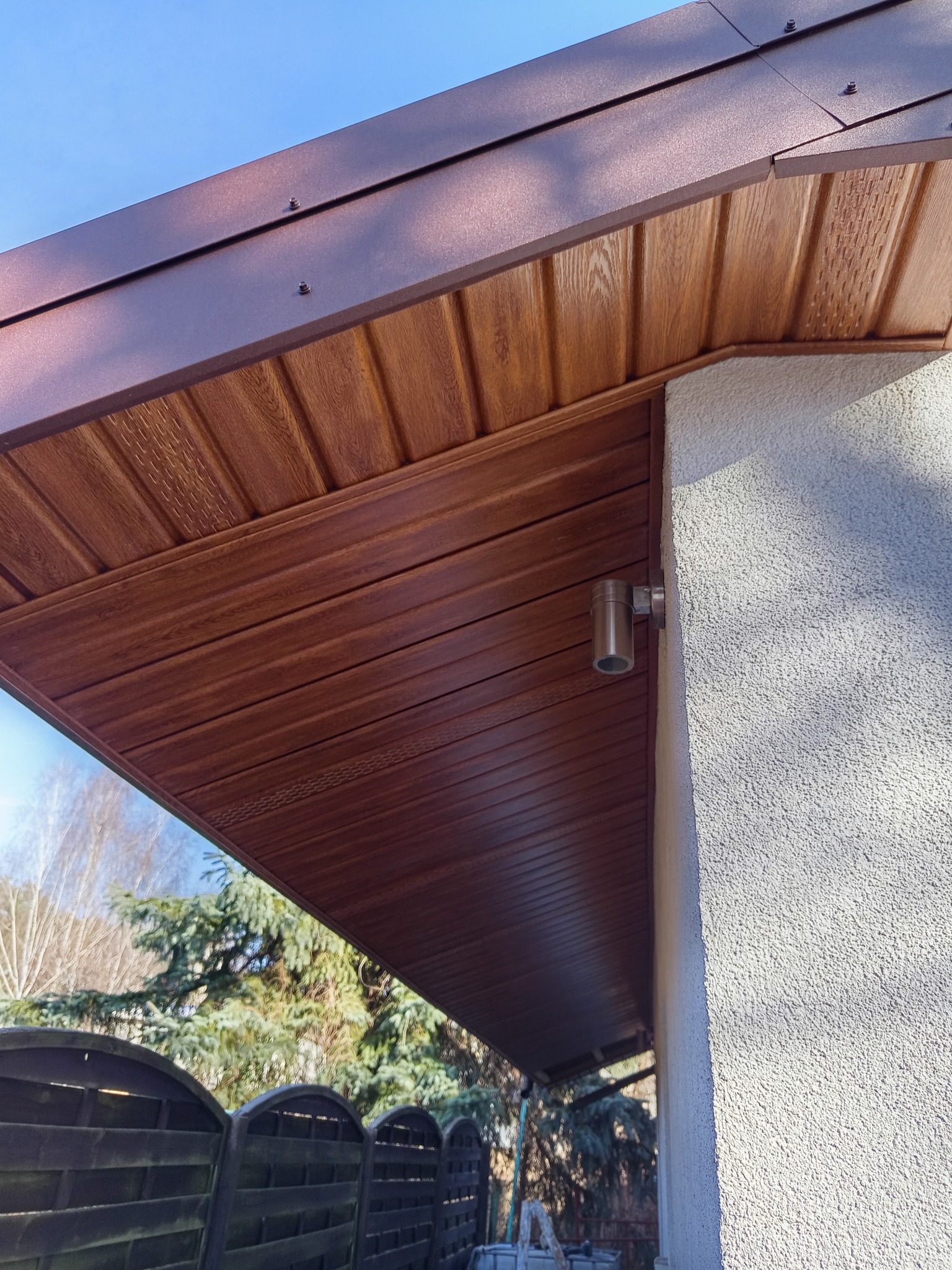 Podbitka dachowa z paneli drewnopodobnych w kolorze brązowym, zamontowana przy ścianie budynku z oświetleniem zewnętrznym. Widok z dołu na tle zieleni.
