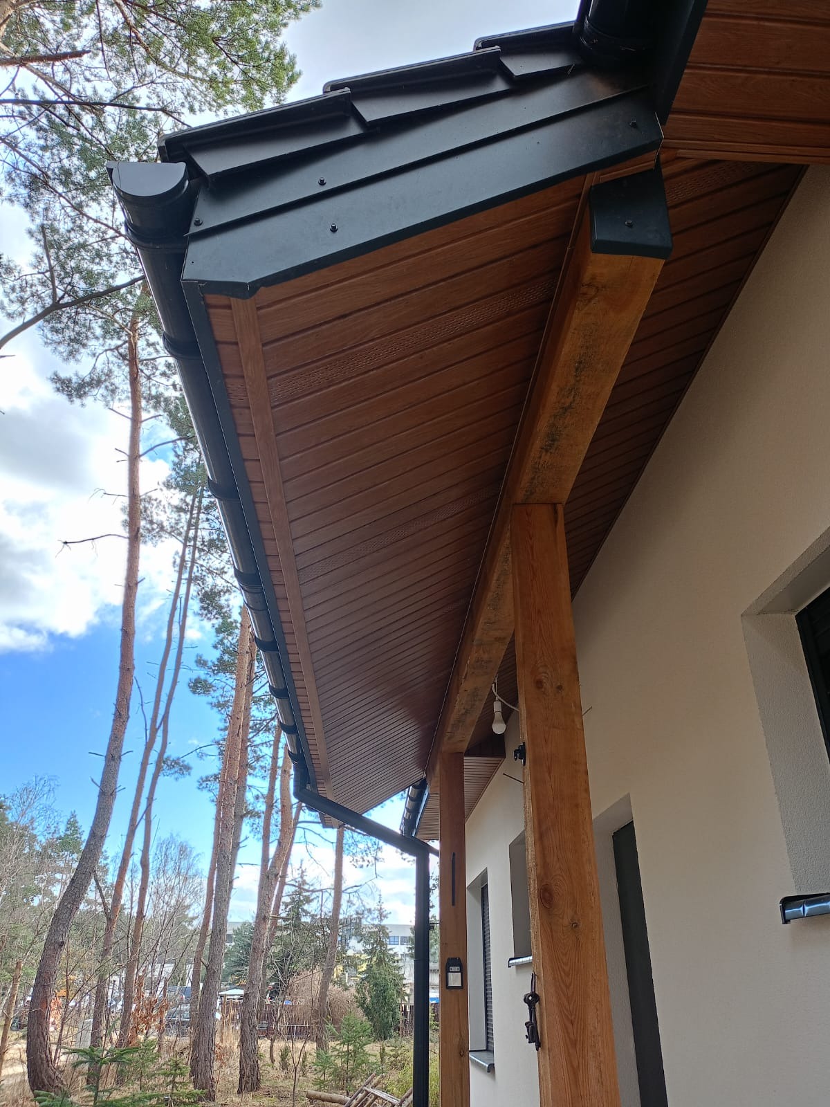 Drewniana podbitka dachowa z czarnymi rynnami, widok z dołu na tle błękitnego nieba i drzew. Elementy wykończeniowe elewacji domu jednorodzinnego.