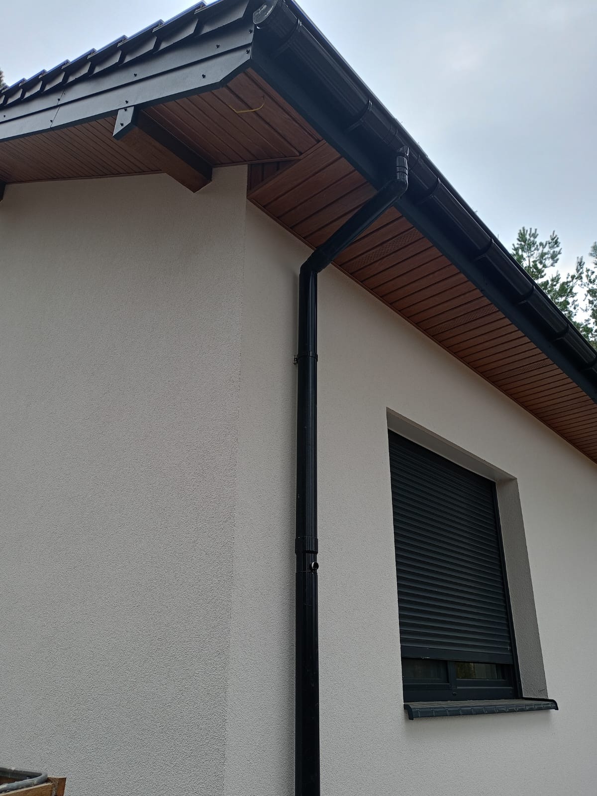 Elewacja domu jednorodzinnego w Toruniu z zamontowanym systemem rynnowym w kolorze czarnym i oknem z roletą zewnętrzną. Widoczna drewniana podbitka dachowa.