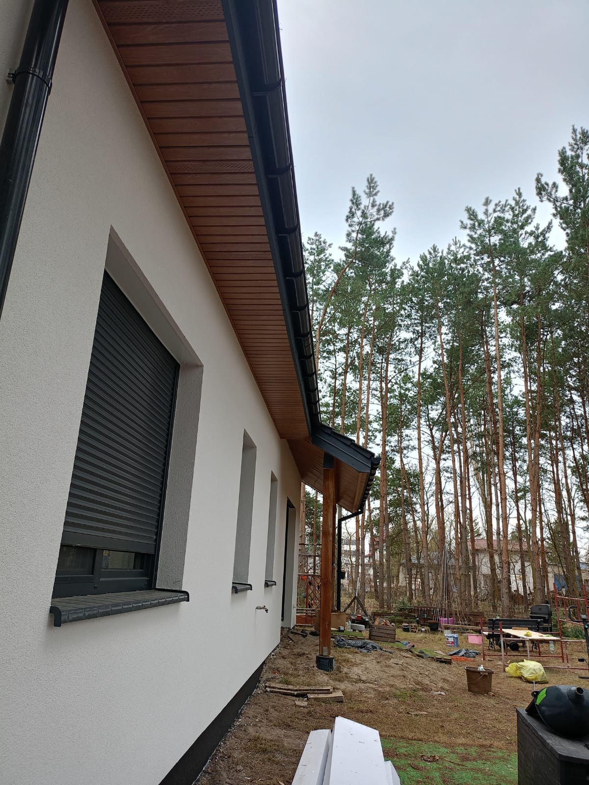 Biała elewacja domu z szarymi roletami i drewnianą podbitką, widok z boku na tle lasu. Świeżo otynkowana ściana z widocznymi detalami wykończenia.