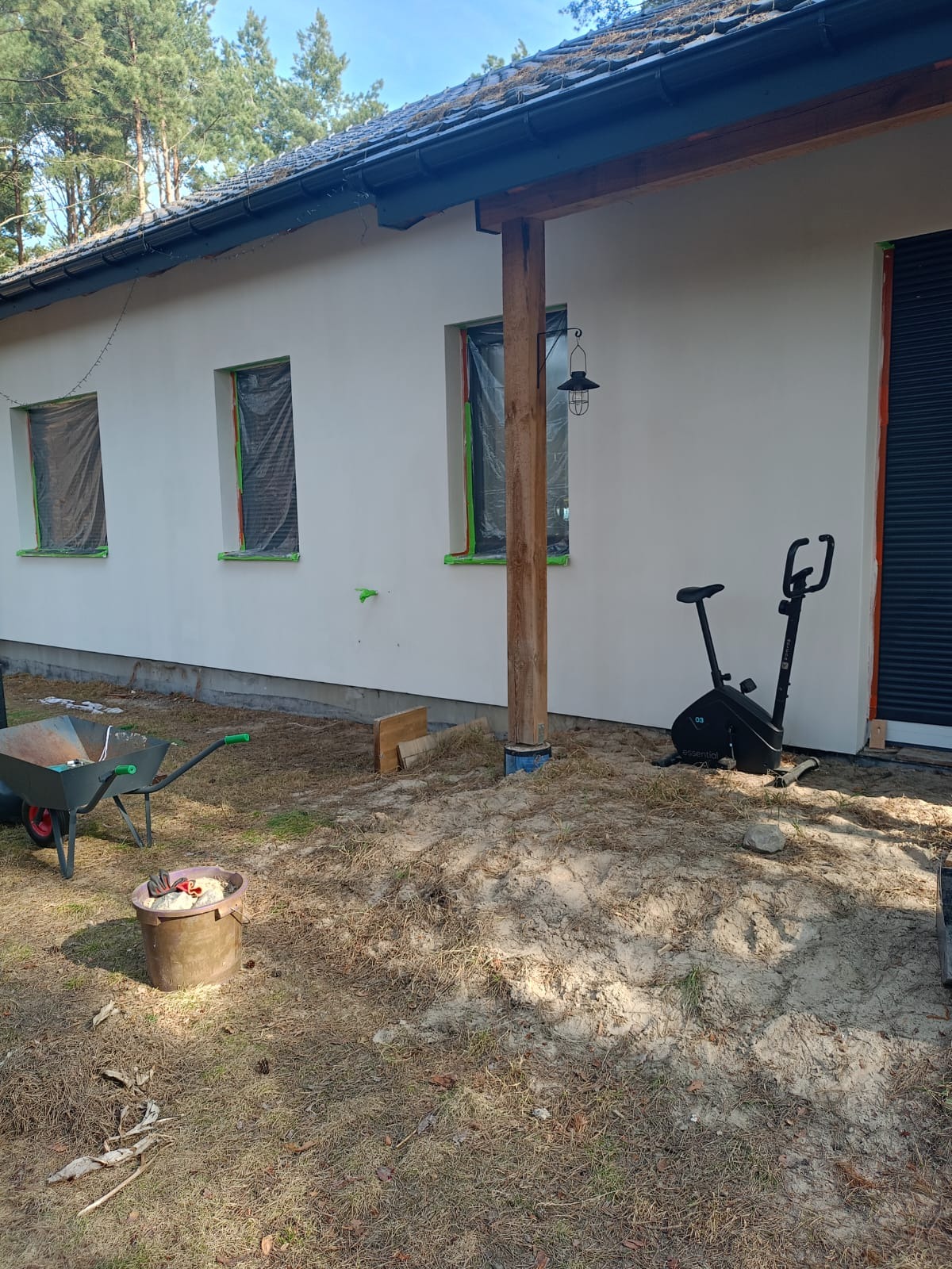 Biała elewacja domu po tynkowaniu, okna zabezpieczone folią, widoczny rower stacjonarny i taczka z narzędziami. Ujęcie z boku budynku, w tle las.
