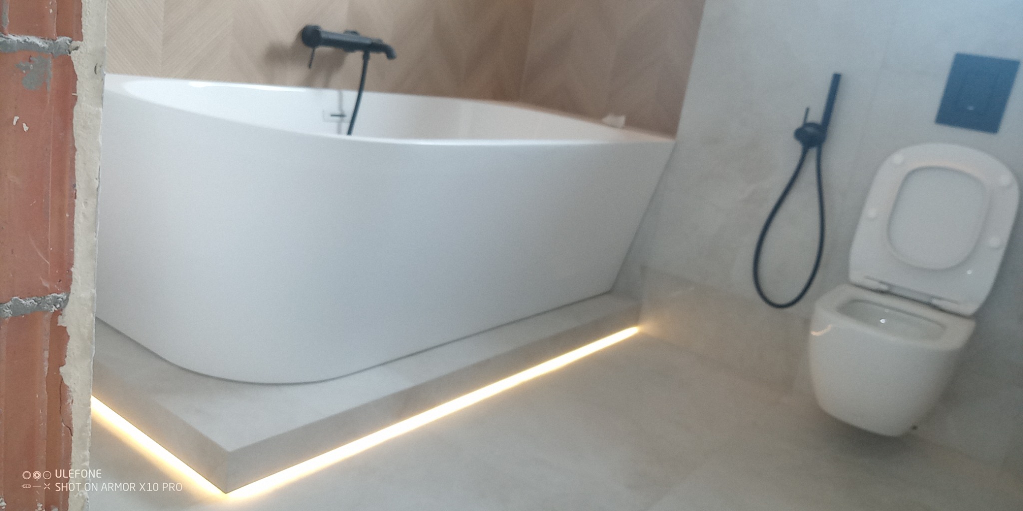 Nowoczesna łazienka po remoncie: biała wanna z podświetleniem LED, wisząca toaleta z czarną armaturą. Fragment ściany z surowej cegły dodaje kontrastu.