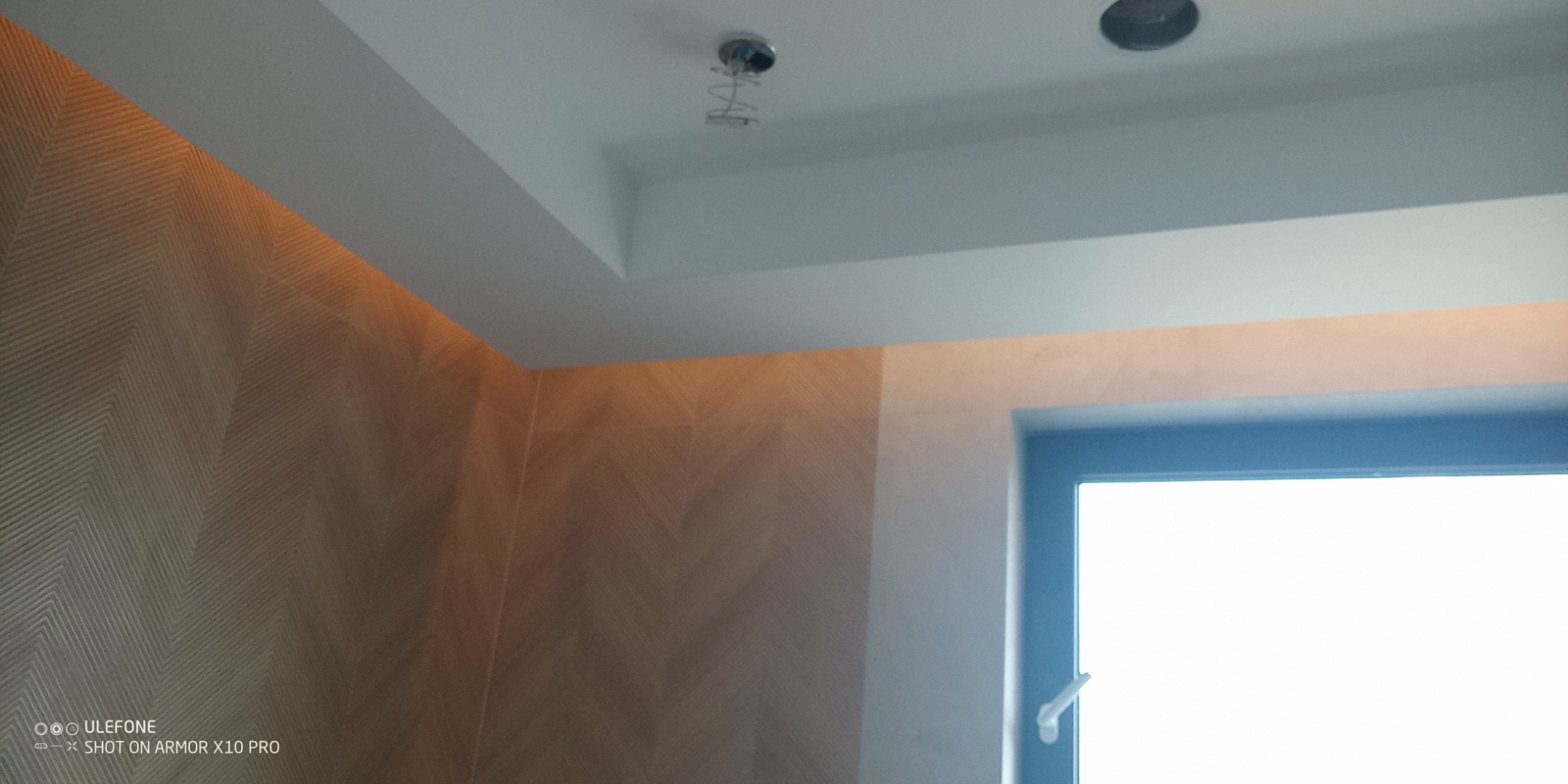 Wykończone wnętrze: ściana z wzorem jodełki, podwieszany sufit z oświetleniem LED i otworem na lampę. Widok na okno. Neutralna kolorystyka.