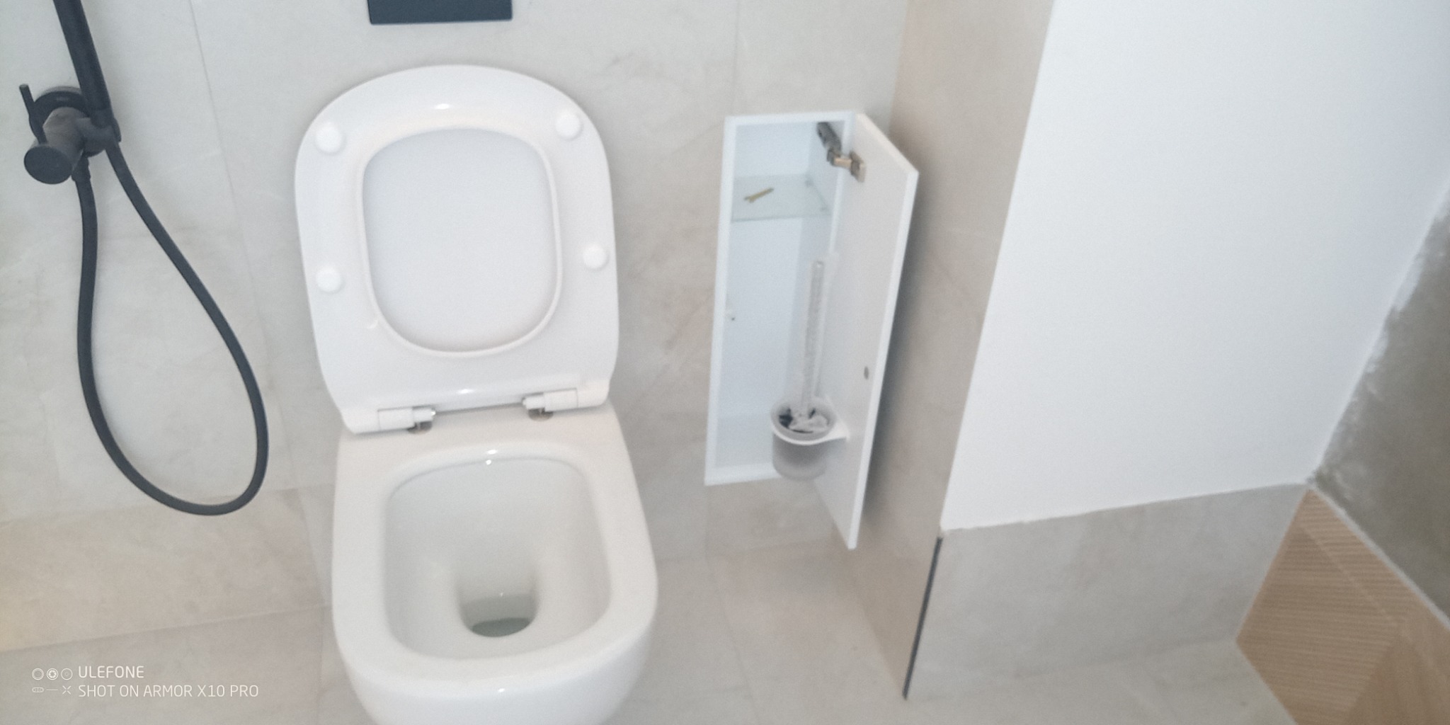 Biała toaleta z otwartą klapą i czarną słuchawką prysznicową na tle beżowych płytek. Obok szafka z szczotką do WC. Minimalistyczny design łazienki po remoncie.