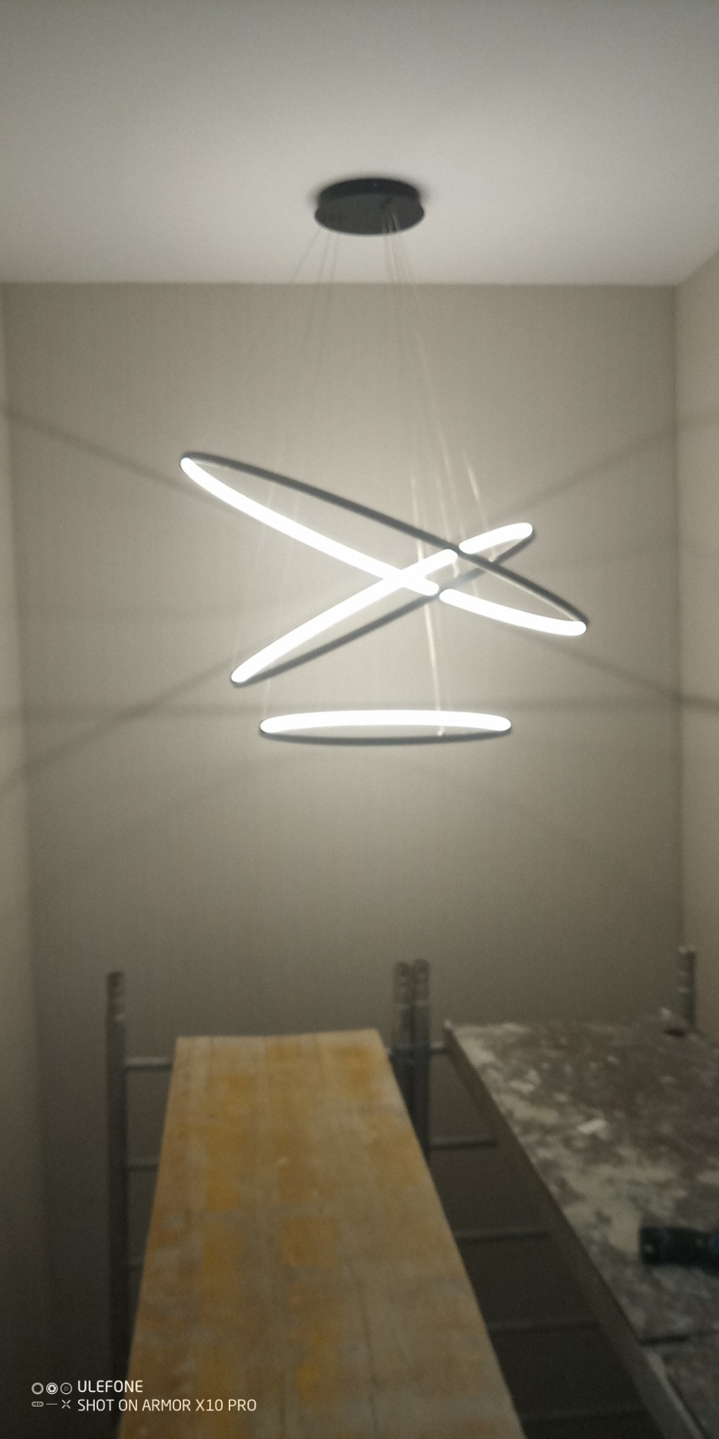 Nowoczesna lampa wisząca z trzema okręgami LED w surowym wnętrzu. Widoczne rusztowania malarskie, co sugeruje prace wykończeniowe. Minimalistyczny design oświetlenia.