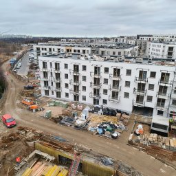 Maciej Wilczak - Wysoka perspektywa na plac budowy z widokiem na powstające budynki mieszkalne, materiały budowlane i robotników w odblaskowych kamizelkach. Pochmurne niebo w tle.