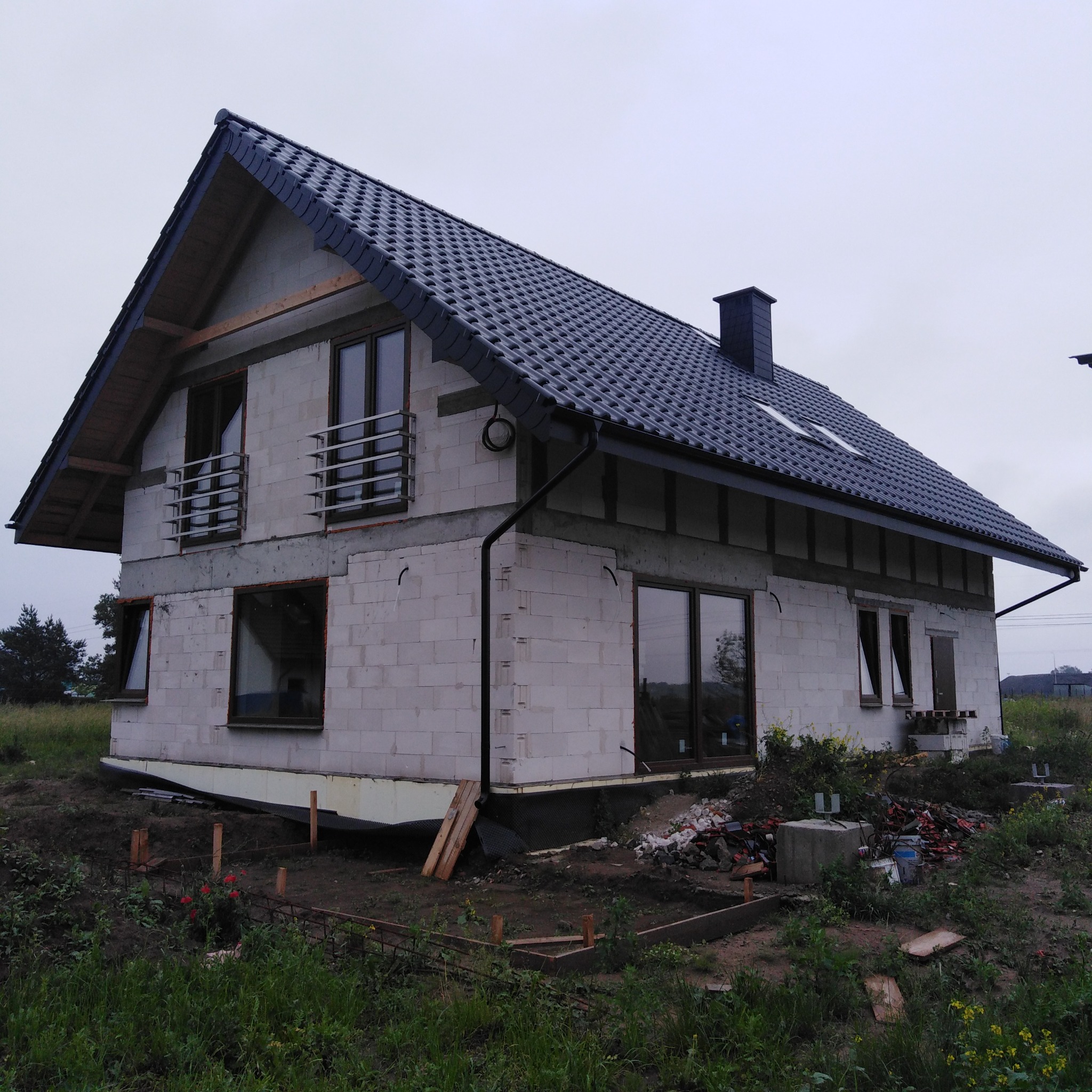 Surowy stan domu z pustaków z ciemną dachówką w Cedrach Małych. Widoczne okna i balkony. Teren wokół w trakcie przygotowania, z elementami konstrukcyjnymi.