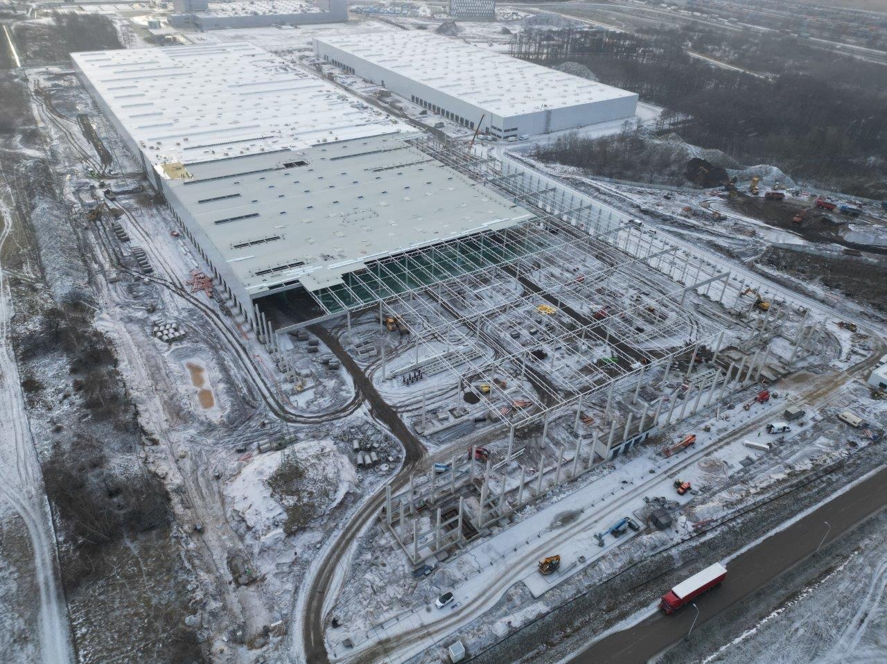 Budowa hali magazynowej w zimowej scenerii z lotu ptaka. Widoczne konstrukcje stalowe, betonowe słupy i częściowo zadaszona powierzchnia. Sprzęt budowlany na ośnieżonym terenie.