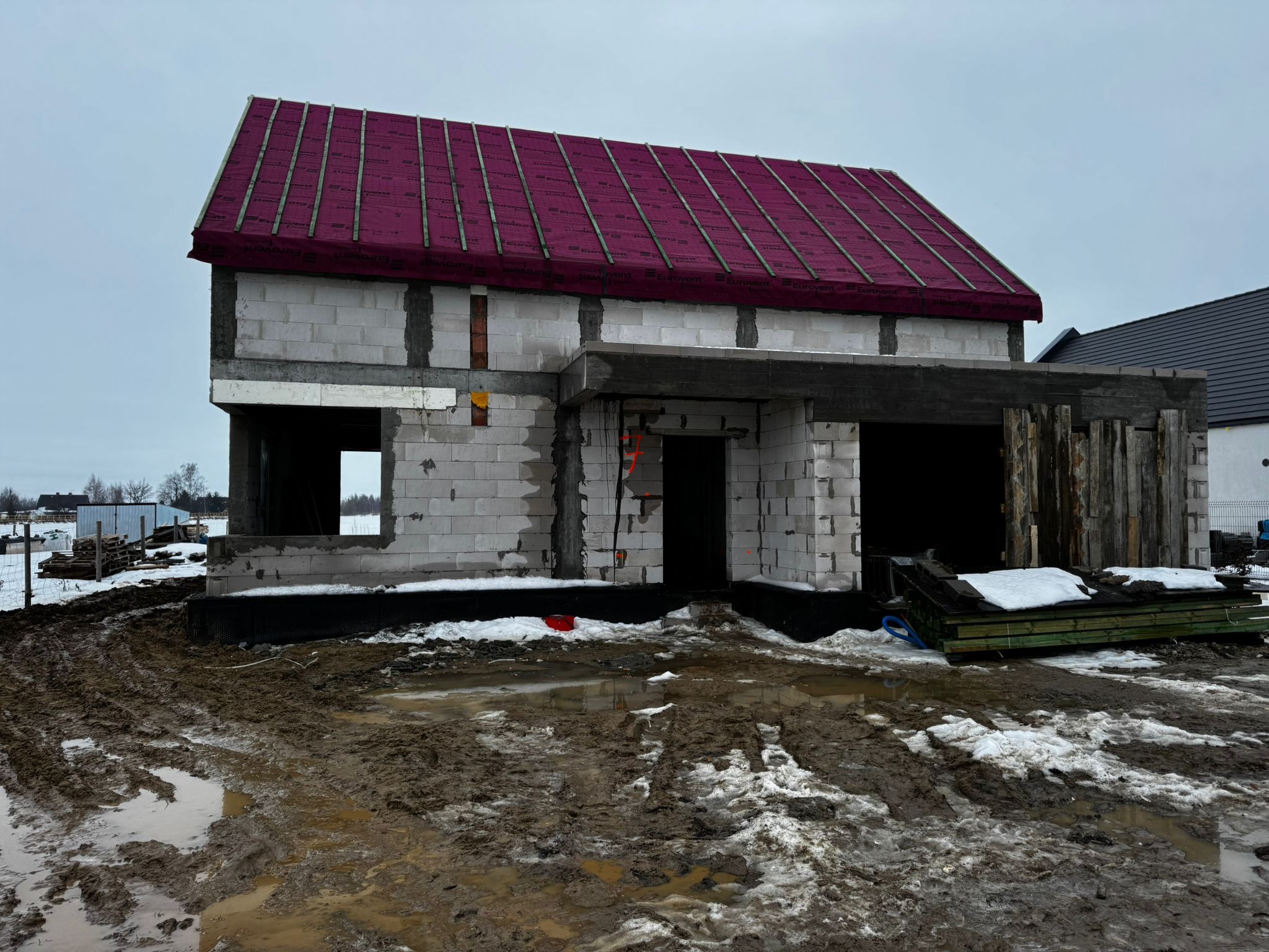 Niedokończony dom z białych bloczków, z czerwonym dachem i widocznymi elementami konstrukcyjnymi. Błotniste podłoże z resztkami śniegu. Surowy stan budynku.