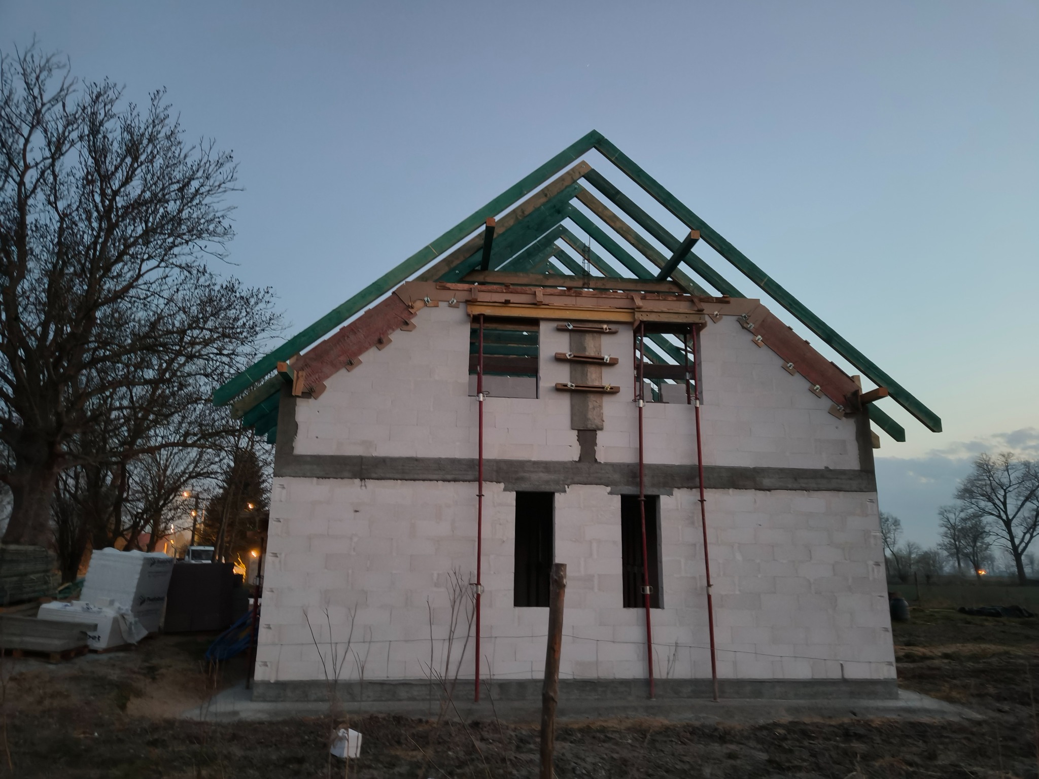 Wznoszenie domu z białych bloczków: ściany z otworami okiennymi, drewniana konstrukcja dachu w trakcie budowy, podpory pionowe. Widok z zewnątrz, w tle drzewa i niebo o zmierzchu.