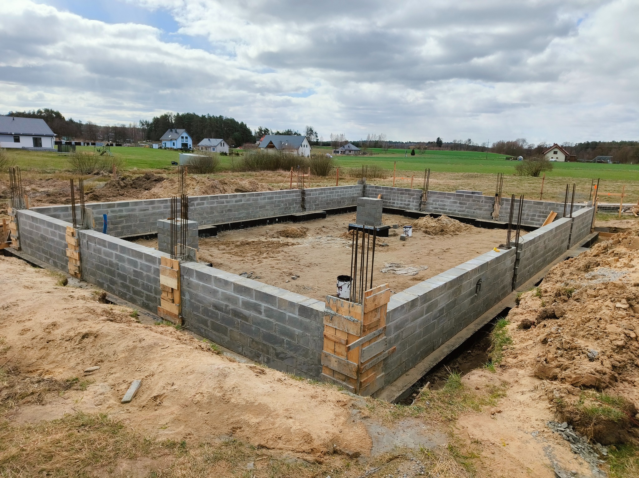 Wzniesione ściany fundamentowe z bloczków betonowych, zbrojenie pionowe, szalunki drewniane w narożnikach. Widok na plac budowy i okoliczne domy w tle.