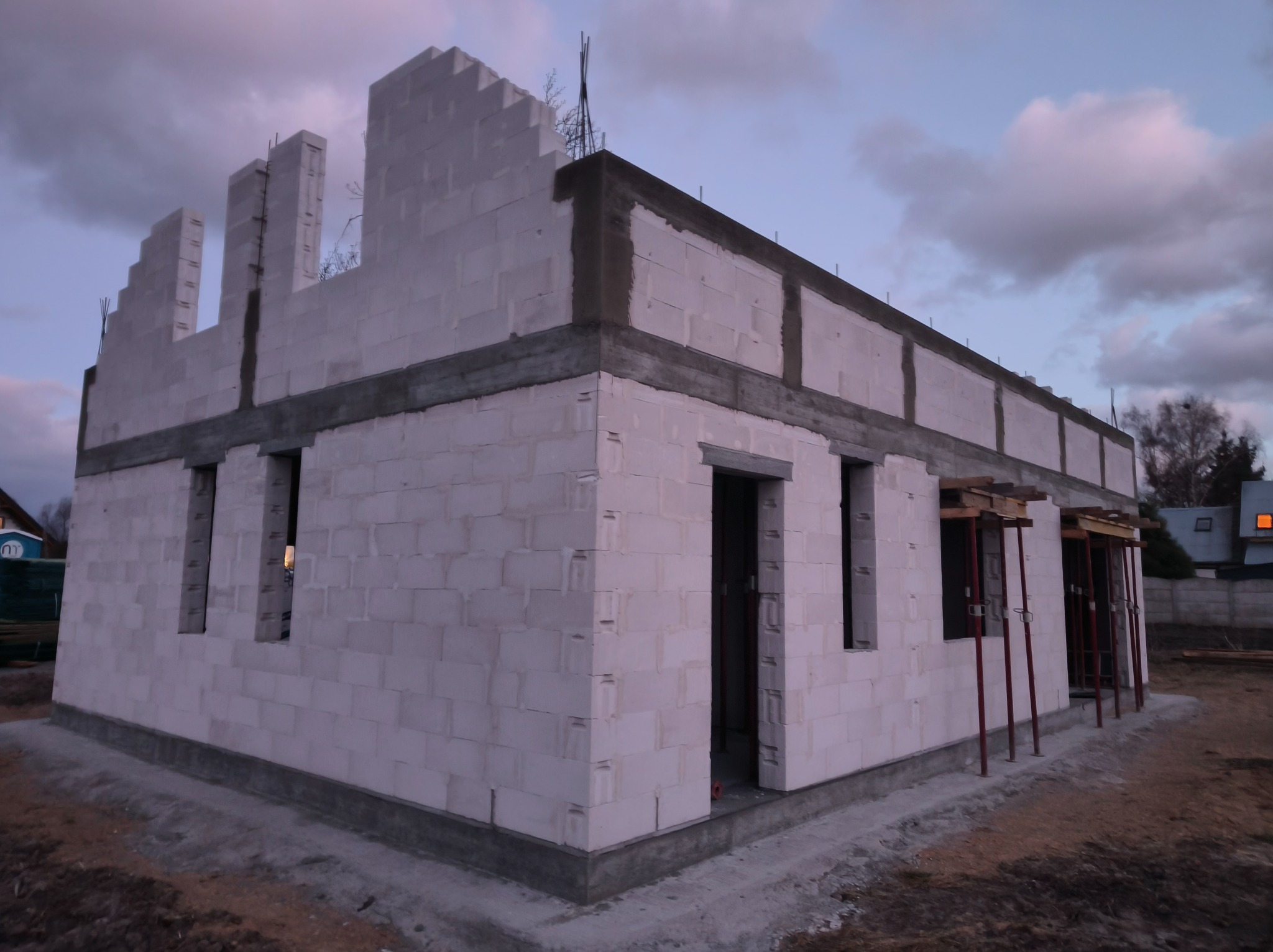 Budowa domu z betonu komórkowego w trakcie realizacji. Widoczne ściany, otwory okienne i drzwiowe, oraz podpory szalunkowe. Ujęcie z zewnątrz, wczesny etap budowy.
