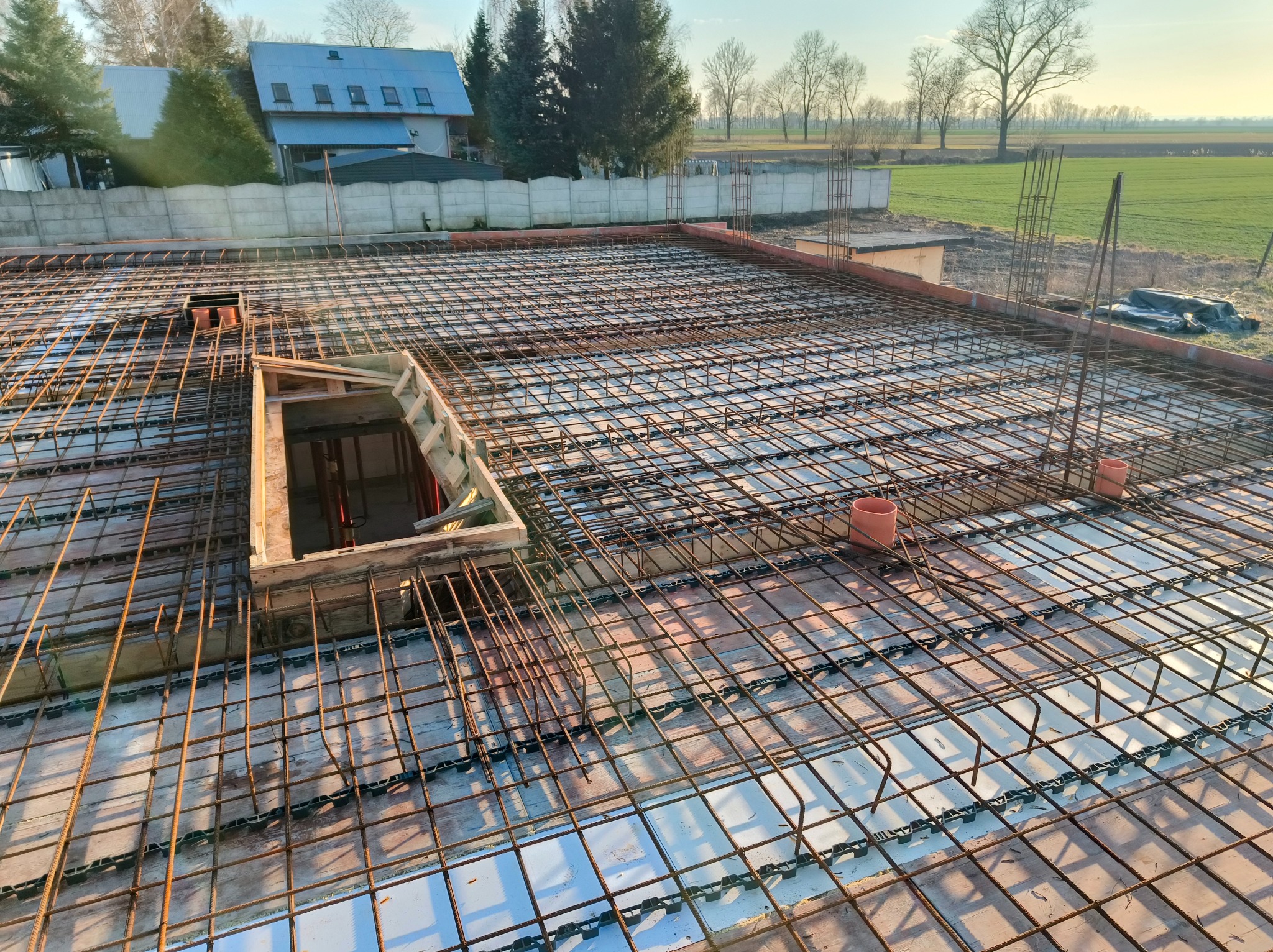 Zbrojenie stropu budynku w trakcie budowy, widok z góry na siatkę z prętów stalowych, szalunek otworu, w tle dom i pole. Słoneczny dzień.