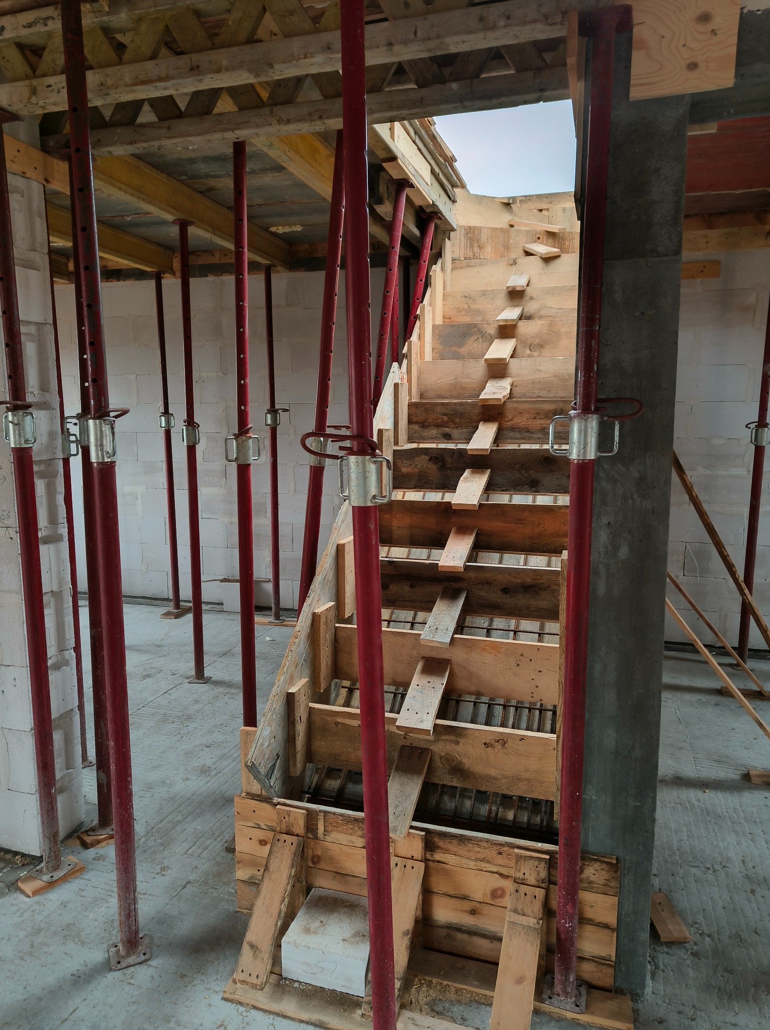 Szczegółowe ujęcie drewnianego szalunku schodów na budowie, wspartego czerwonymi stemplami. Widoczne zbrojenie i betonowy filar. Surowy stan prac.