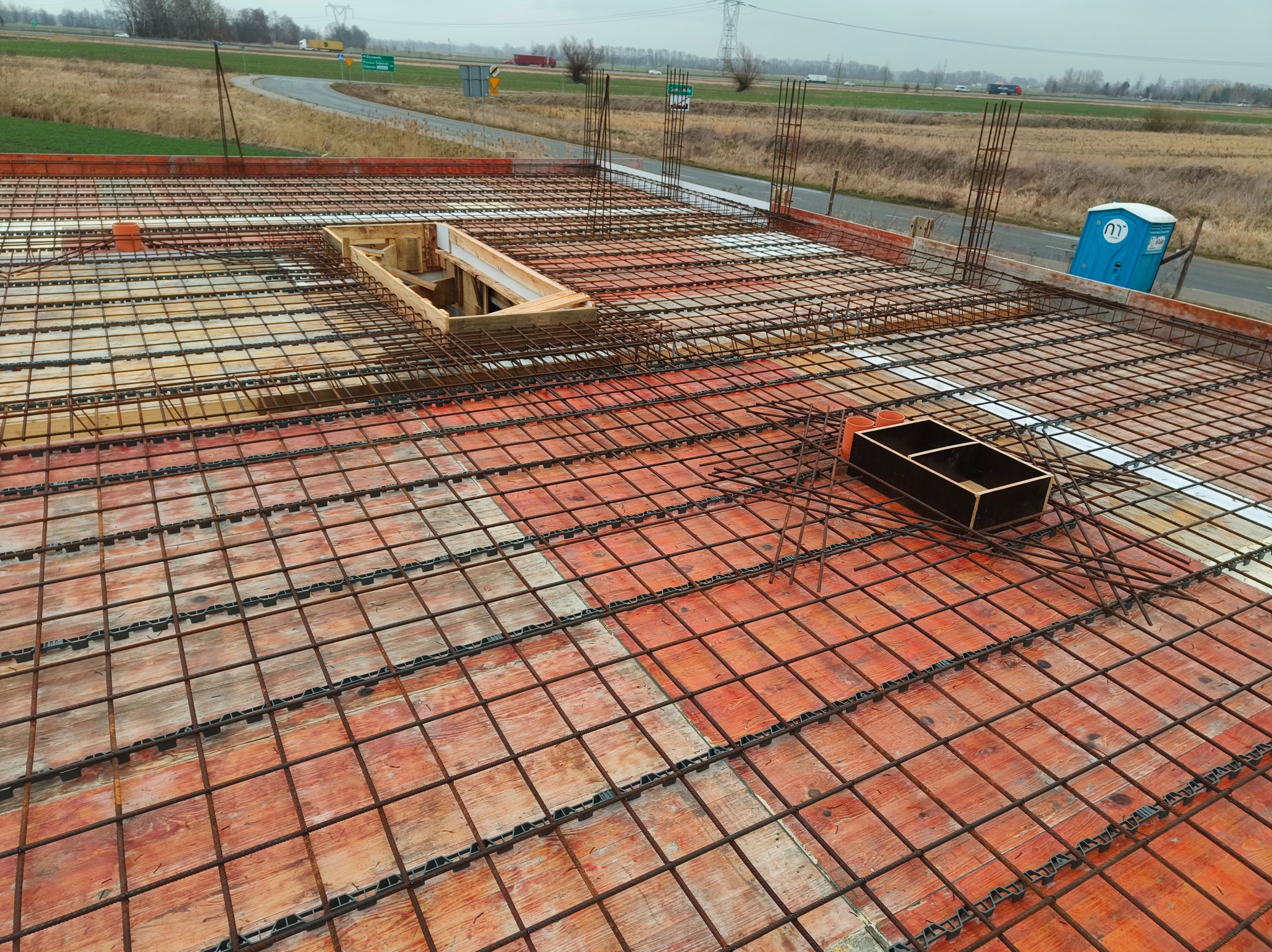 Zbrojenie stropu budynku w trakcie budowy, widok z góry na siatkę z prętów stalowych i szalunki. W tle krajobraz z drogą i polem.