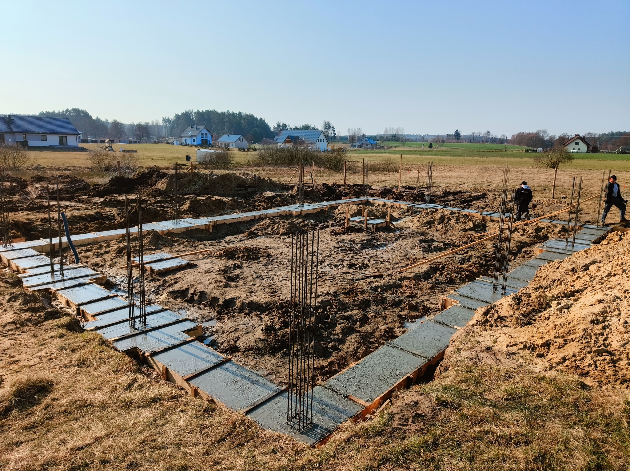 Wylane fundamenty pod dom jednorodzinny z wystającymi prętami zbrojeniowymi, widoczne szalunki. W tle zabudowa wiejska i dwóch pracowników budowlanych.