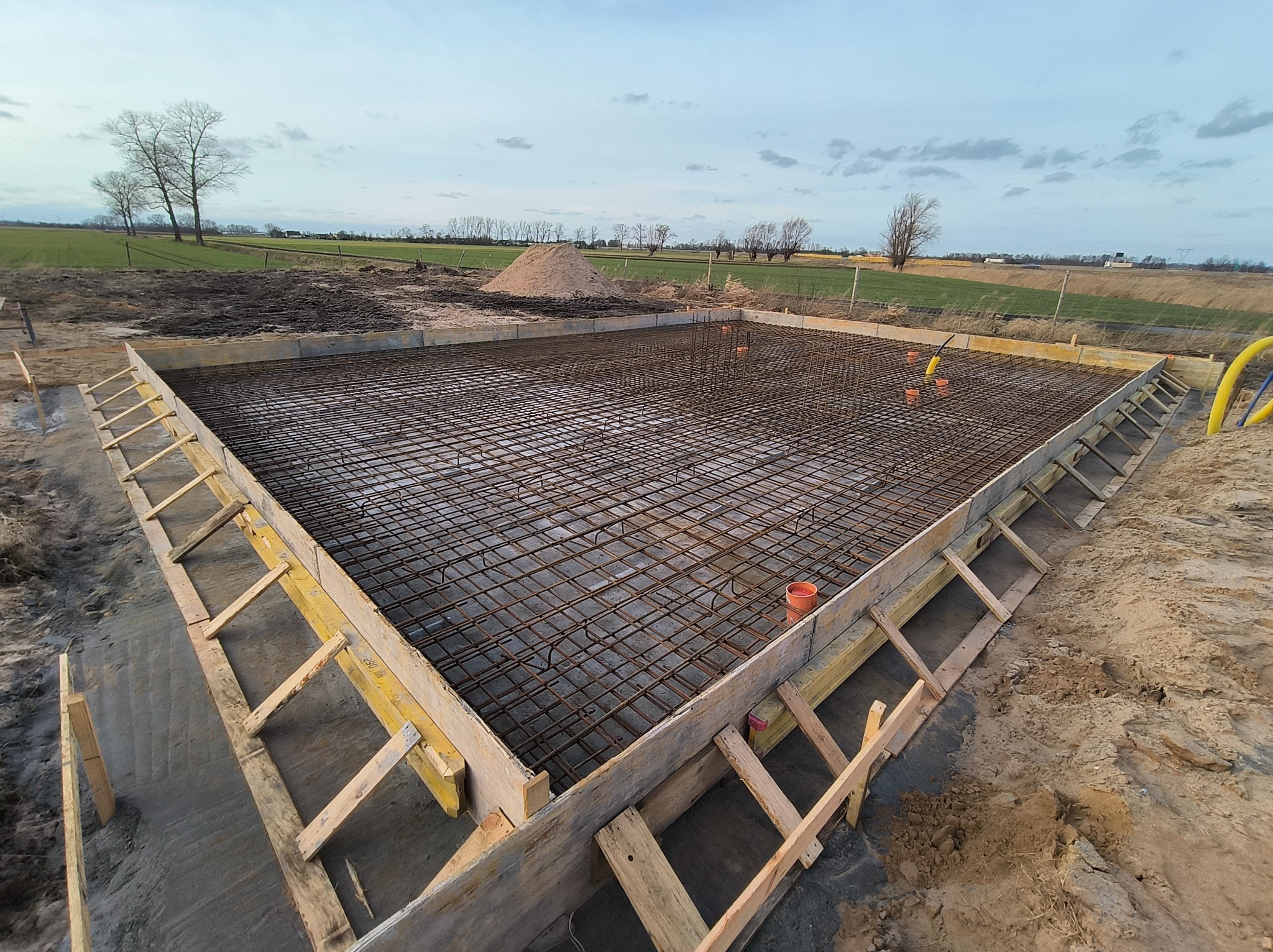 Zbrojenie fundamentów w drewnianym szalunku, widok z góry. Na drugim planie ziemia, trawa i drzewa. Widoczne pomarańczowe rury wewnątrz konstrukcji.