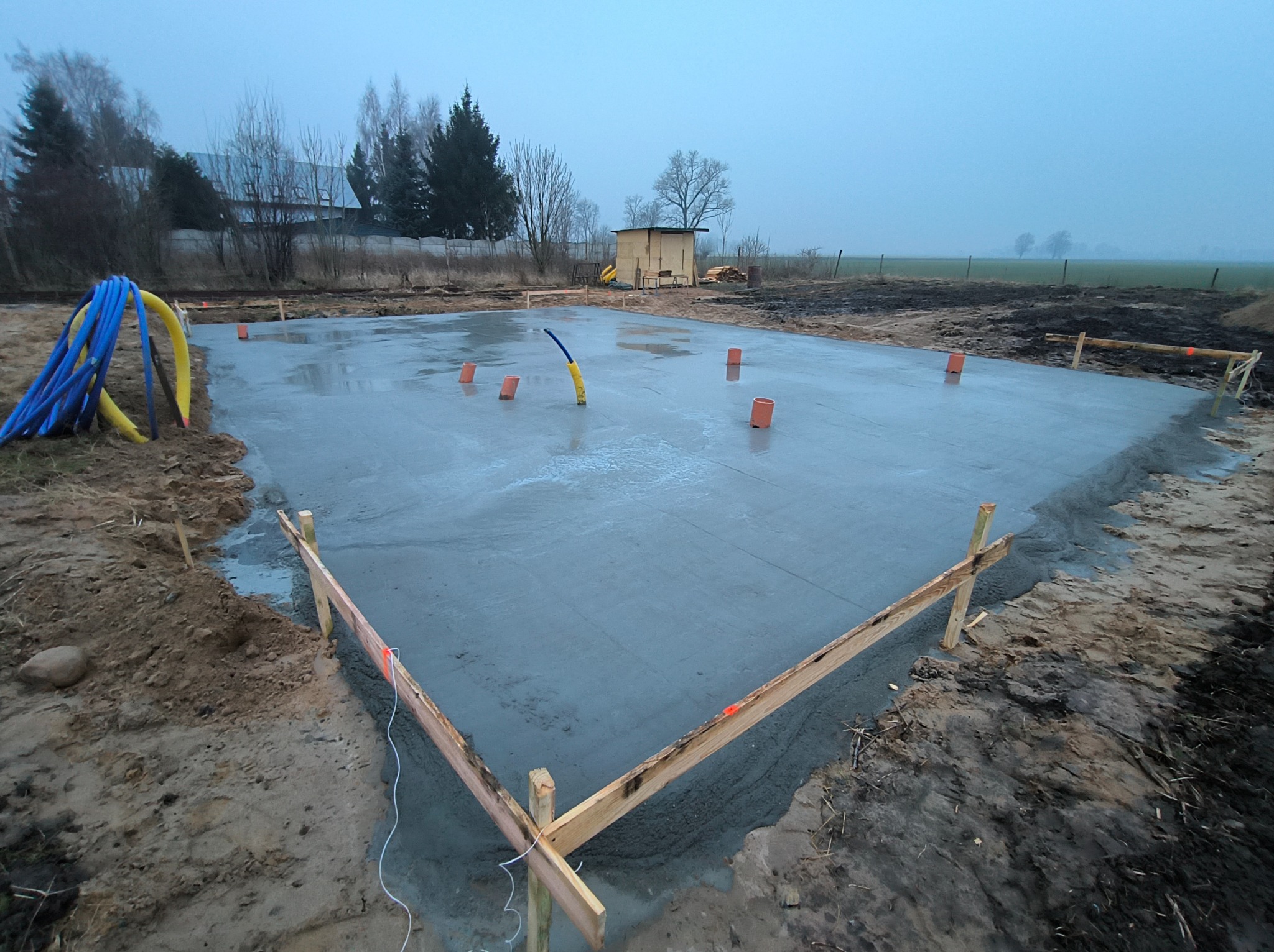Świeżo wylana, gładka płyta fundamentowa z widocznymi rurami instalacyjnymi i drewnianym szalunkiem, na tle pochmurnego krajobrazu z budynkiem gospodarczym.