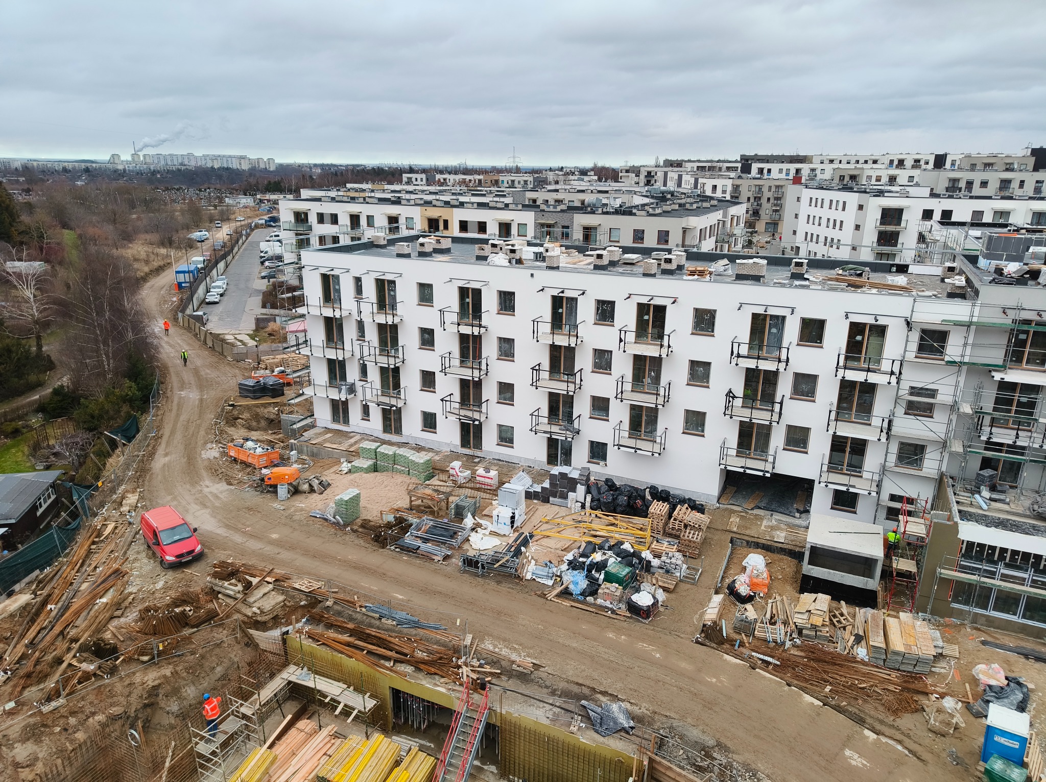 Wysoka perspektywa na plac budowy z widokiem na powstające budynki mieszkalne, materiały budowlane i robotników w odblaskowych kamizelkach. Pochmurne niebo w tle.