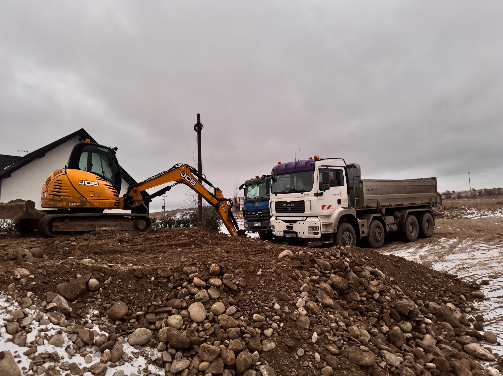 Żółta koparka JCB i biała ciężarówka MAN na placu budowy z ziemią i kamieniami. W tle budynek z ciemnym dachem i pochmurne niebo.