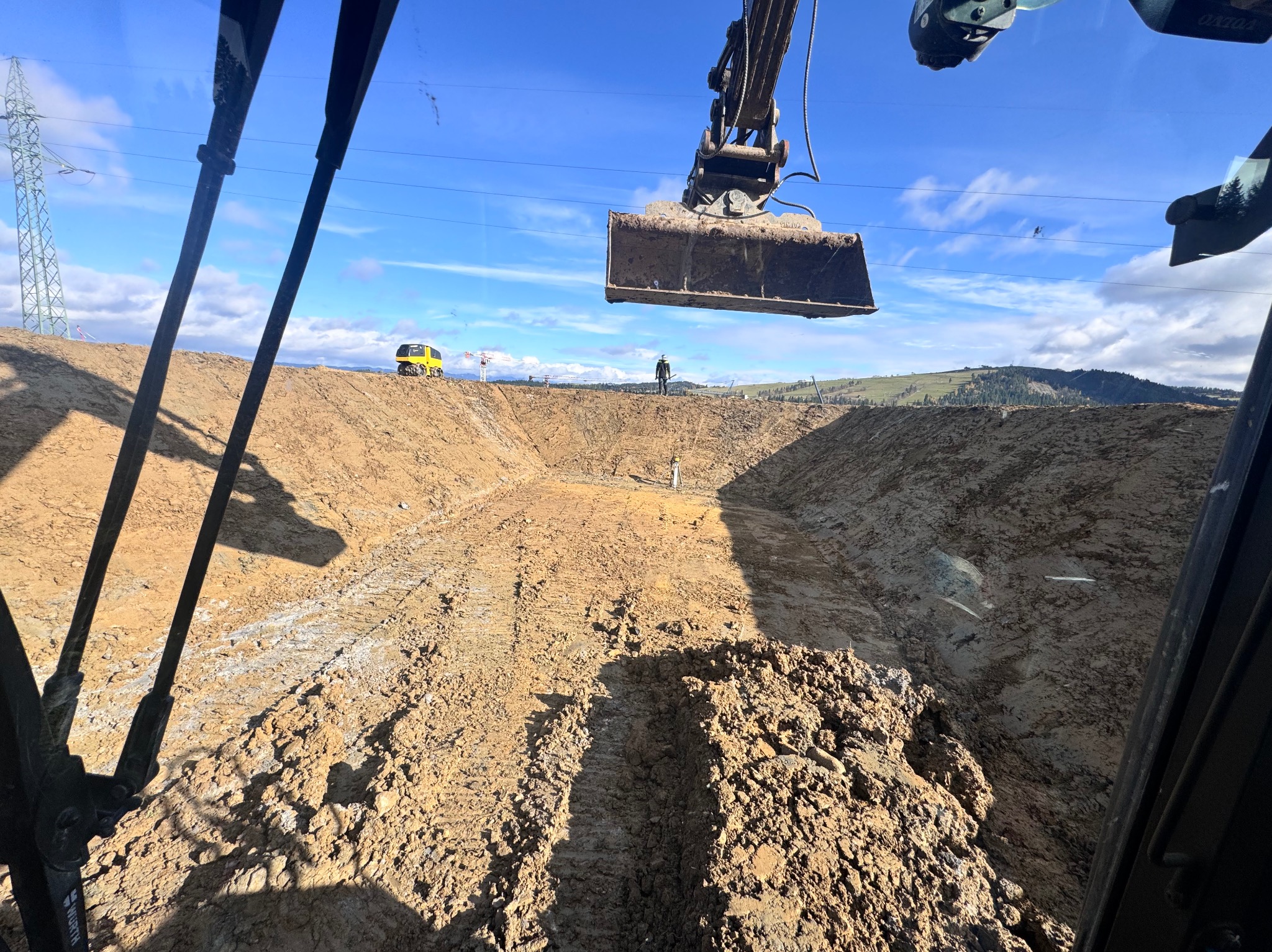 Głęboki wykop ziemny z pracującą koparką w tle, widok z kabiny operatora. Na horyzoncie widoczny pracownik w kasku i żółta maszyna budowlana.