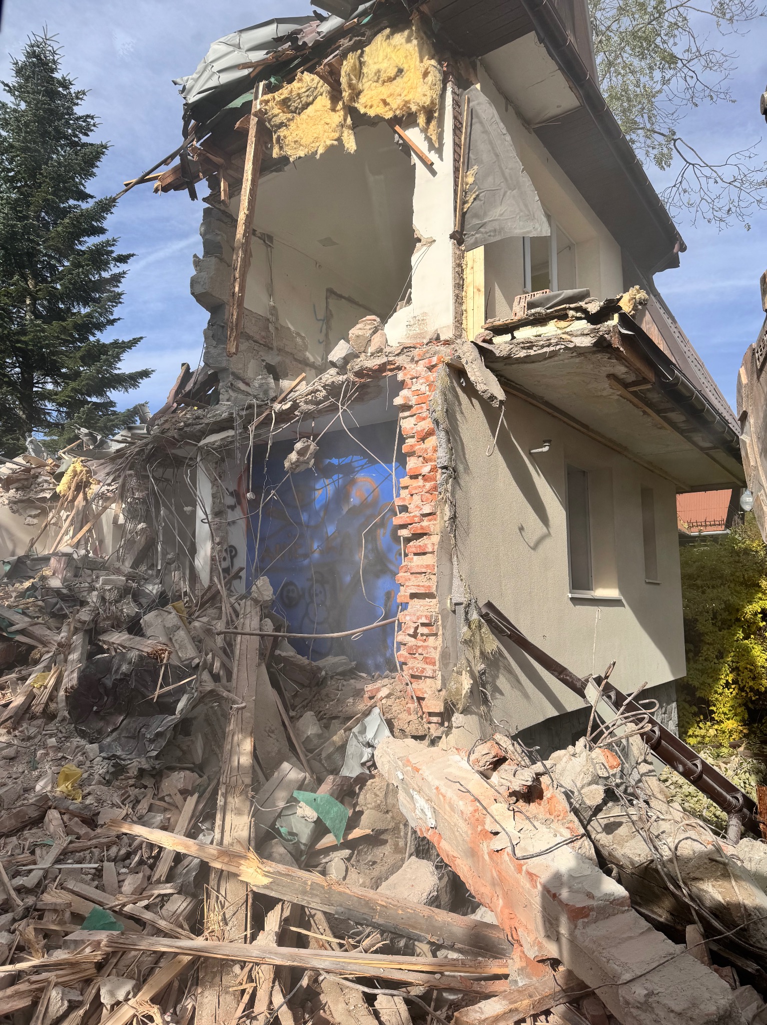 Zrujnowany budynek po rozbiórce, z widocznymi fragmentami ścian, cegieł i elementów konstrukcyjnych. W tle drzewo iglaste i fragment nieba.