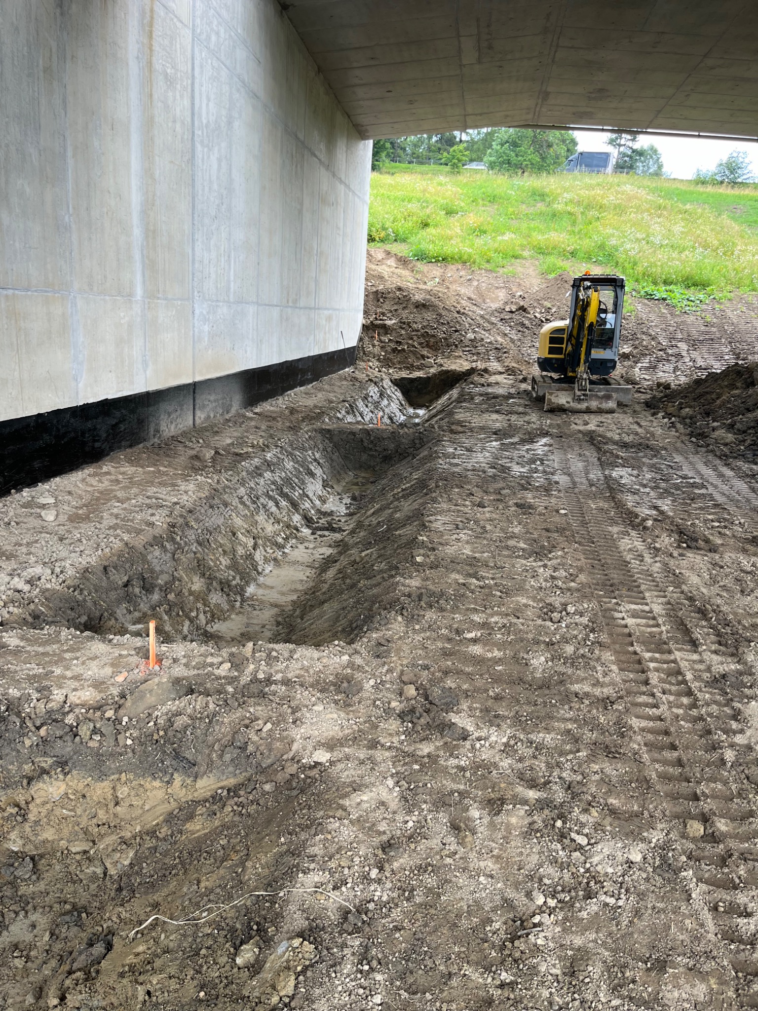 Żółta minikoparka przy wykopie wzdłuż betonowego fundamentu. Widoczne ślady opon na ziemi i zielone wzgórze w tle. Prace ziemne pod konstrukcją.