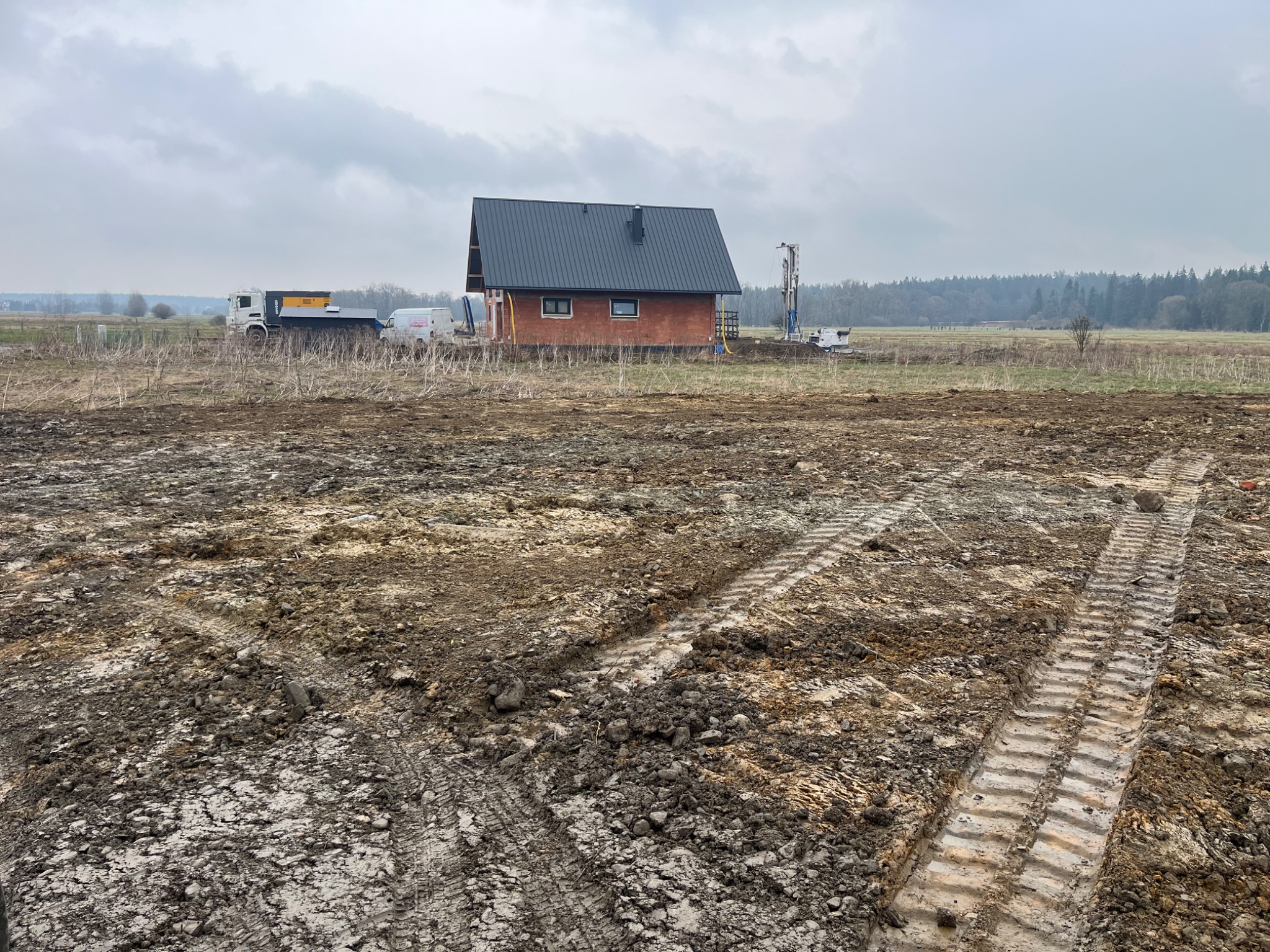 Budowa domu w stanie surowym otoczona rozjeżdżonym terenem. W tle widoczny sprzęt budowlany i transportowy. Pochmurne niebo.