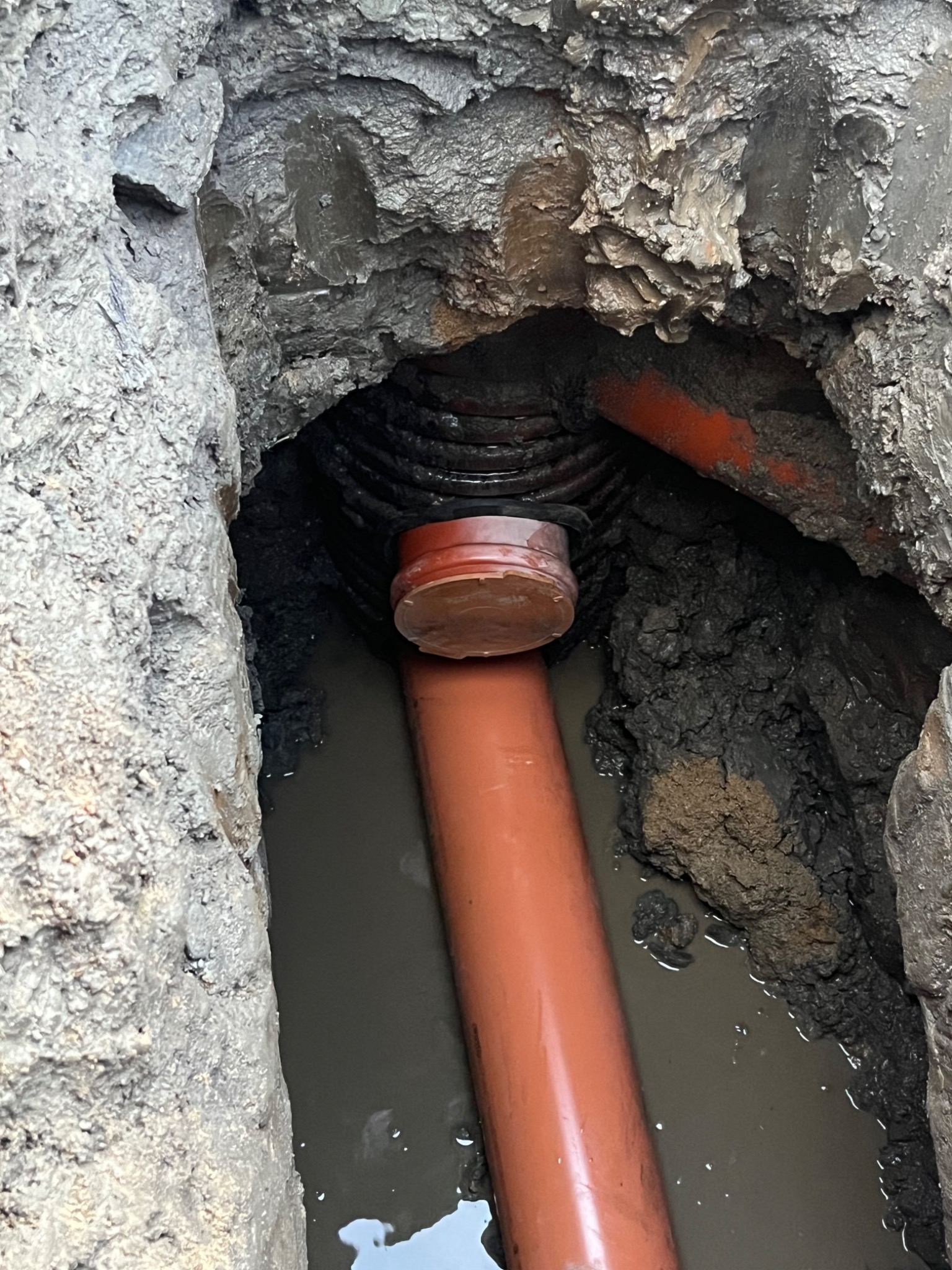 Pomarańczowa rura kanalizacyjna w wykopie wypełnionym wodą, widoczne połączenie rur i struktura ziemi. Ujęcie z góry, blisko rury.