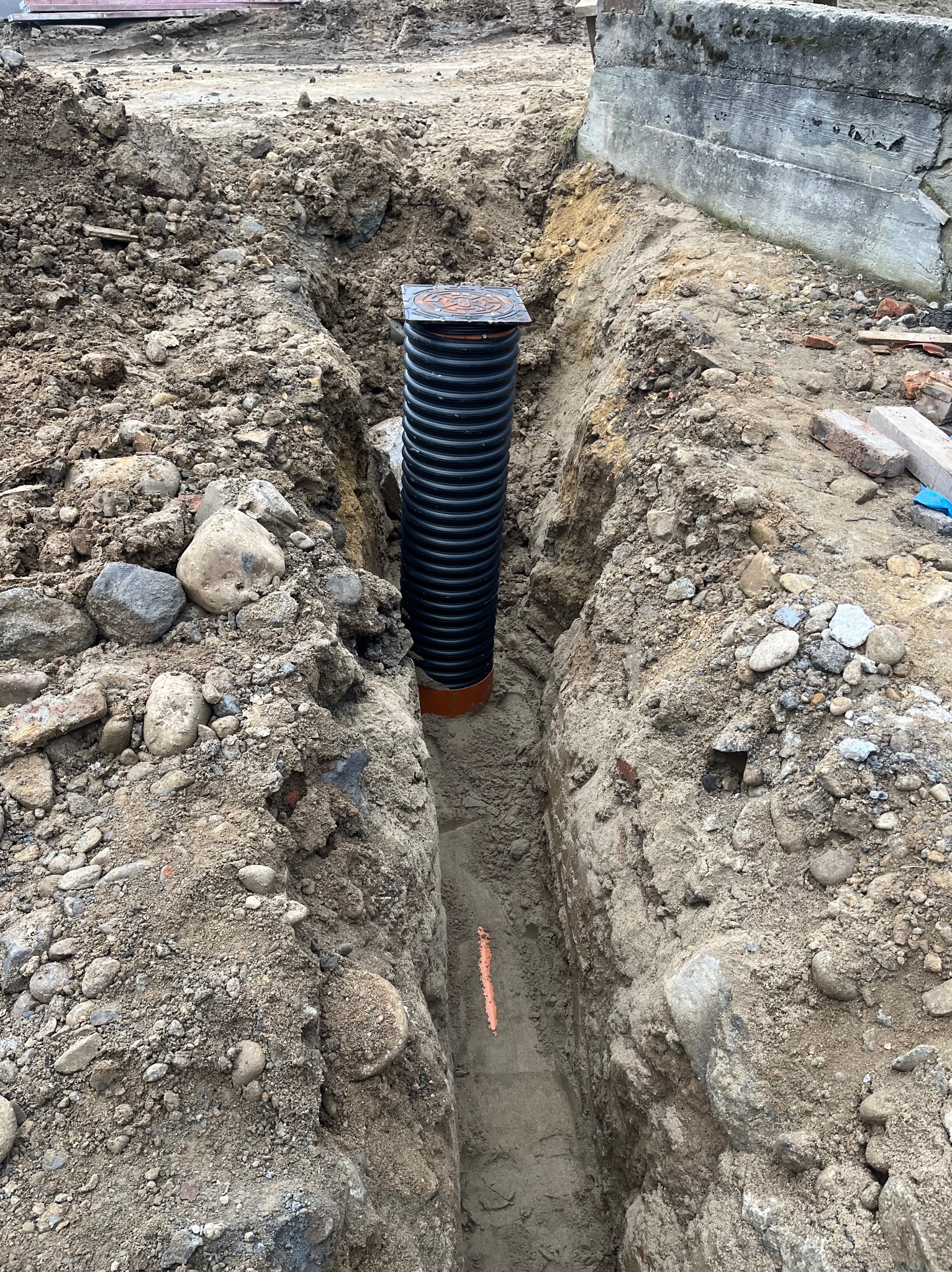 Czarna, karbowana rura drenażowa z pomarańczowym łącznikiem, umieszczona w wąskim wykopie w ziemi z kamieniami, widok z góry.