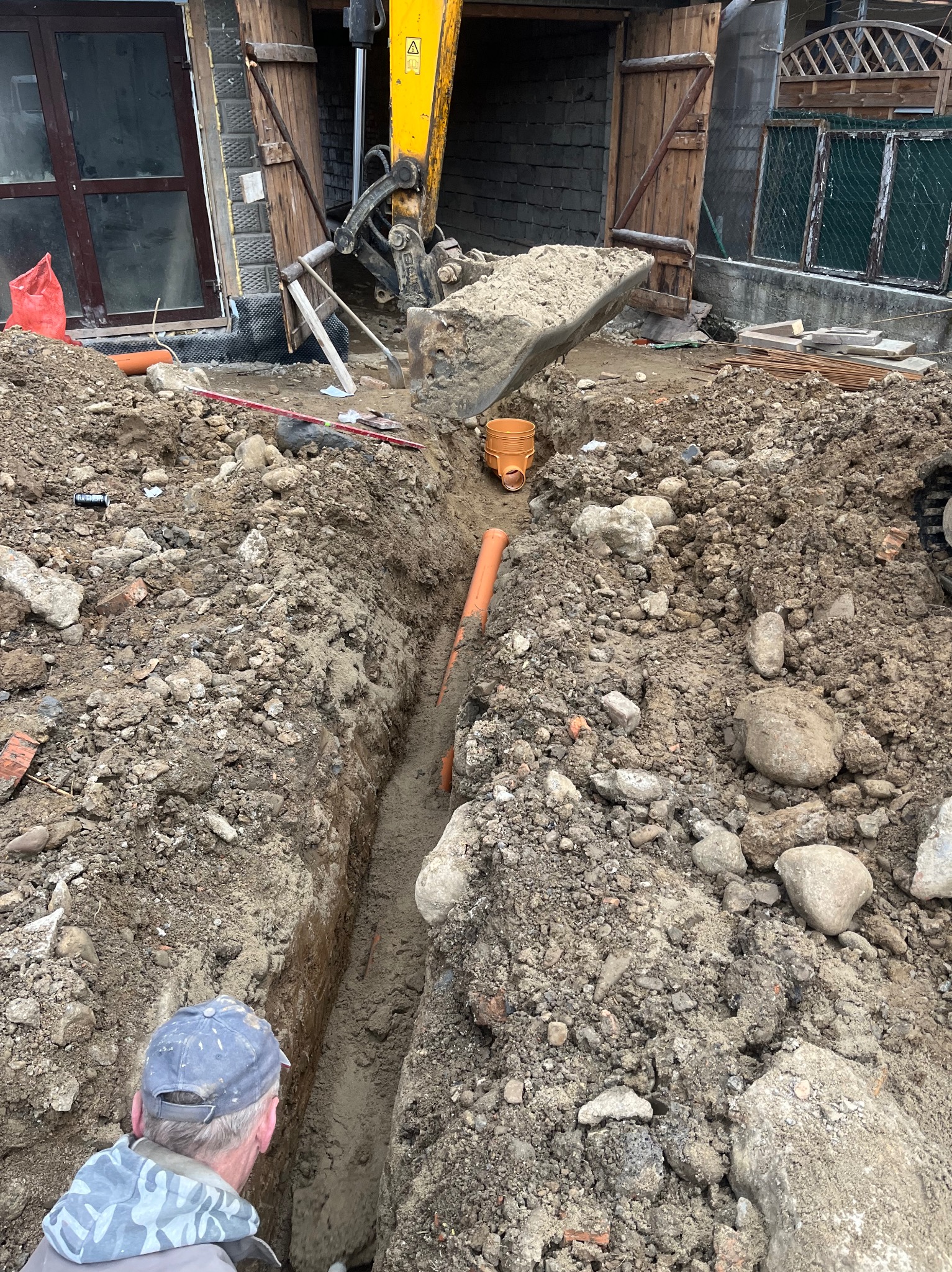 Wąski wykop pod rury drenarskie, z żółtą koparką w tle i fragmentem budynku. Widoczny pracownik w czapce. Ujęcie z góry, podkreślające głębokość wykopu.