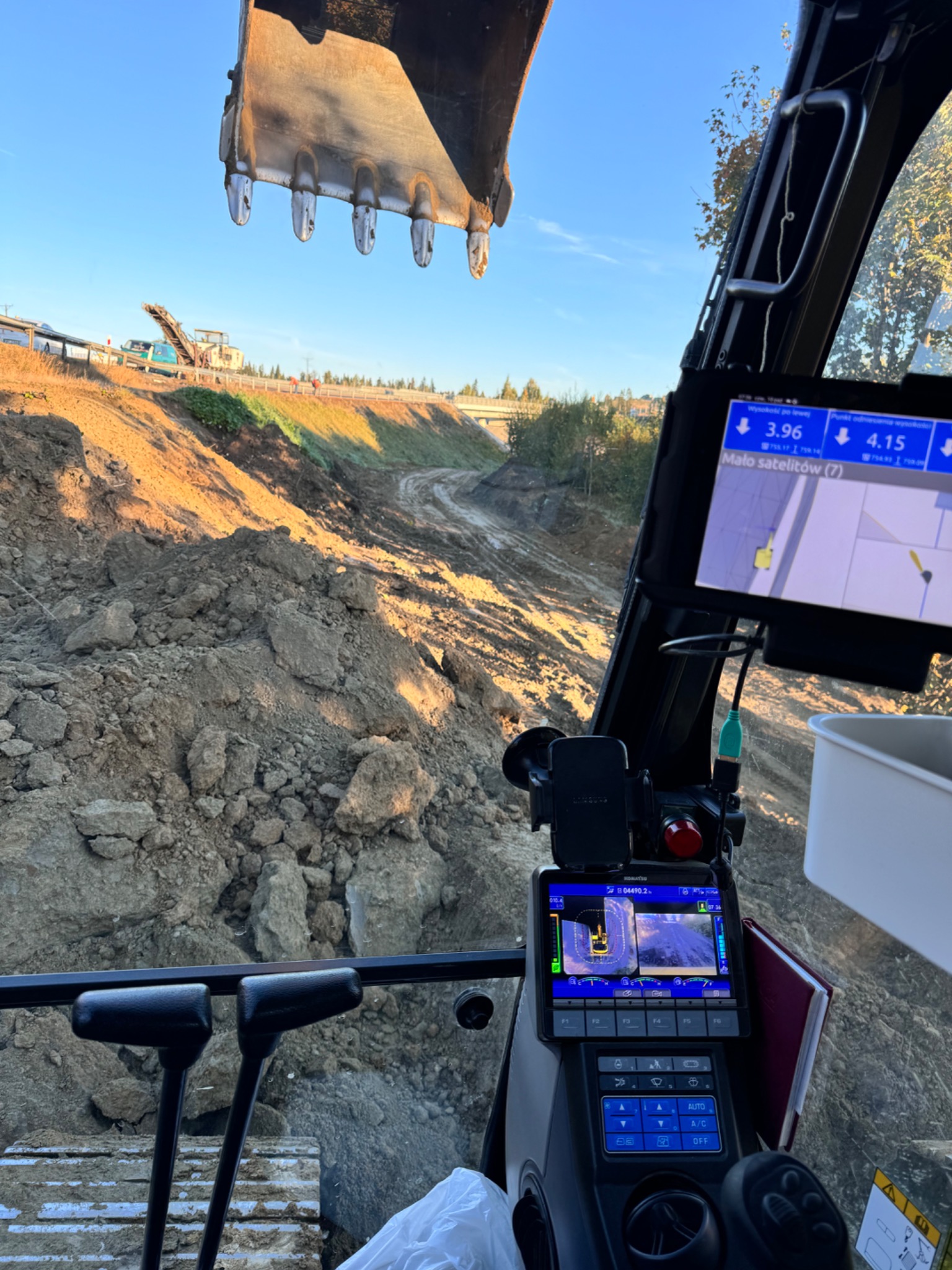 Widok z kabiny koparki na wykopany teren. Na monitorze widać obraz z kamery. W tle widać inny sprzęt budowlany i drzewa. Oświetlenie słoneczne.
