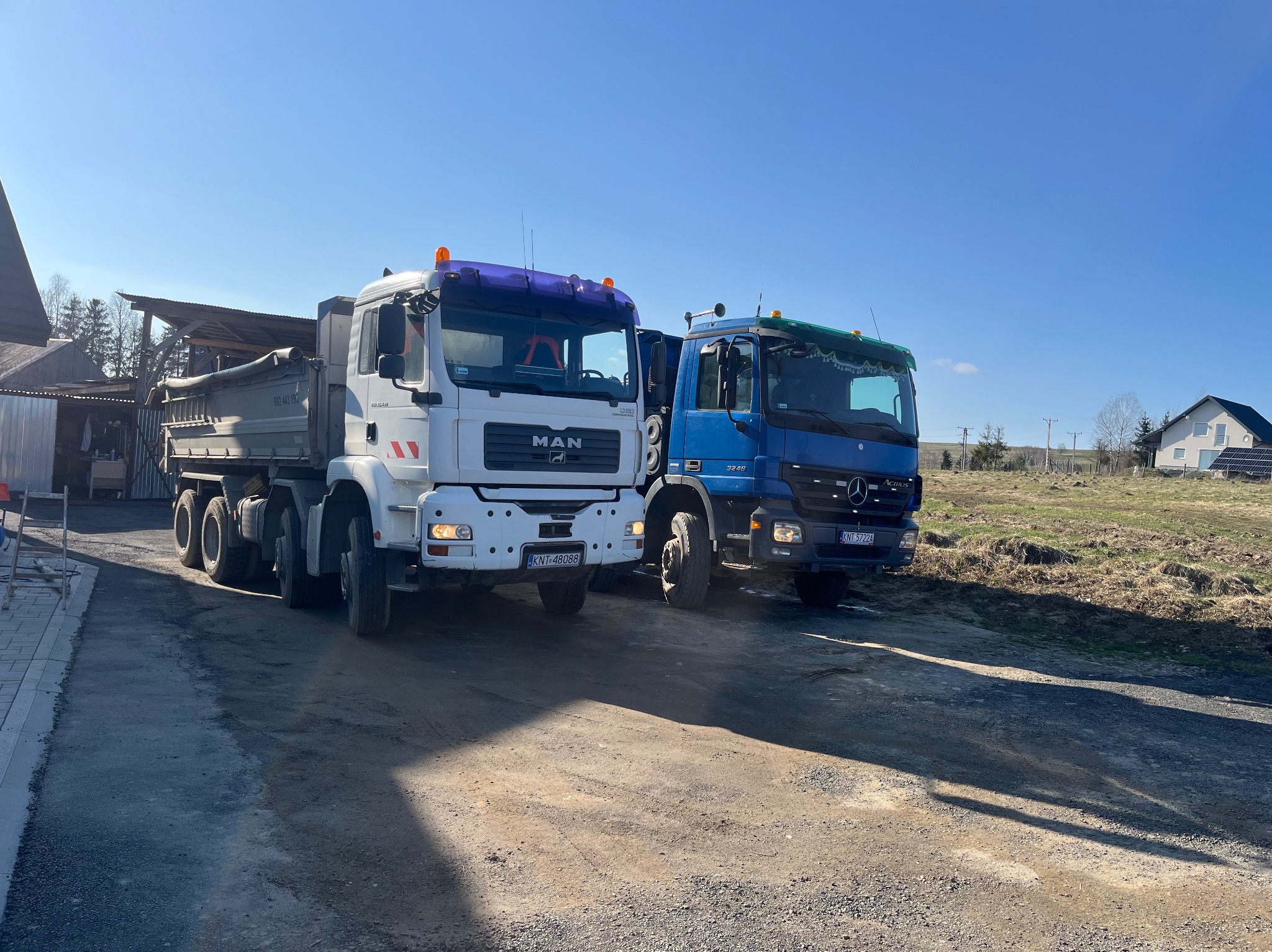 Dwa samochody ciężarowe MAN i Mercedes zaparkowane obok siebie na żwirowej drodze w słoneczny dzień. W tle budynki i pole.