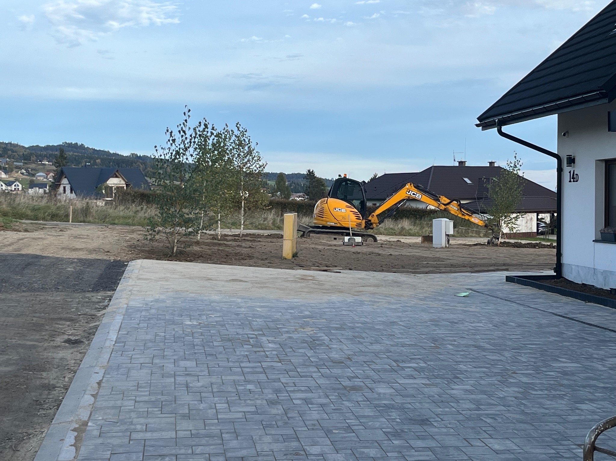 Żółta koparka JCB na działce z kostką brukową, przygotowująca teren pod budowę. W tle domy i drzewa, błękitne niebo.