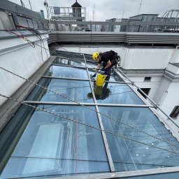 SkyCloud Ruslan Okilka - Pracownik w uprzęży, w żółtym kasku, myje szklany dach budynku z użyciem wiadra i myjki. Widoczne liny zabezpieczające i elementy konstrukcji dachu.