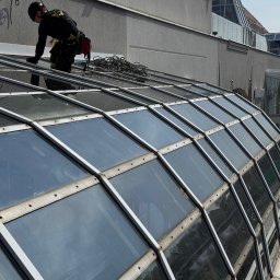 SkyCloud Ruslan Okilka - Pracownik w uprzęży, na szklanym dachu, podczas czyszczenia. Widoczne liny, kask i narzędzia. Ujęcie z dołu, podkreślające wysokość i specyfikę pracy.