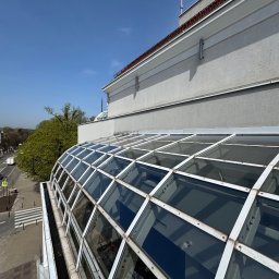 SkyCloud Ruslan Okilka - Szklany dach budynku z widocznymi zabrudzeniami i mchem, ujęcie z góry, fragment elewacji i rynny, błękitne niebo w tle. Widok na ulicę i zieleń.