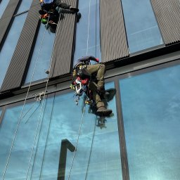 SkyCloud Ruslan Okilka - Dwóch alpinistów przemysłowych na linach czyści szklaną fasadę nowoczesnego biurowca. Widoczne odbicie nieba i słońca w oknach. Ujęcie z dołu.