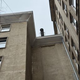 SkyCloud Ruslan Okilka - Ujęcie z dołu na fragment elewacji budynku z rynnami i kominem na dachu. Widoczne okna i szara, chropowata faktura ścian. Pochmurne niebo w tle.