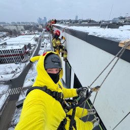 SkyCloud Ruslan Okilka - Osoba w kostiumie Pikachu, w kasku i masce, na linie podczas prac wysokościowych na budynku w zimowej scenerii miejskiej. W tle inni pracownicy i panorama miasta.