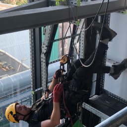 SkyCloud Ruslan Okilka - Pracownik w kasku i uprzęży, zawieszony na linach, wykonuje prace na wysokości na tle konstrukcji stalowej i budynku. Widoczny drugi pracownik w oddali.