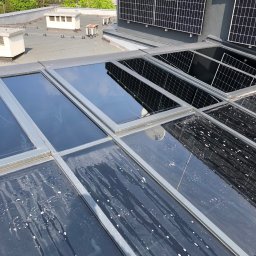 SkyCloud Ruslan Okilka - Zabrudzone okna dachowe na płaskim dachu z panelami słonecznymi w tle. Widoczne smugi i plamy po deszczu. Ujęcie z góry, w słoneczny dzień.