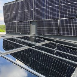 SkyCloud Ruslan Okilka - Szary dach budynku z panelami słonecznymi i świetlikami, w których odbija się niebo. Widok z góry, w tle zieleń drzew.